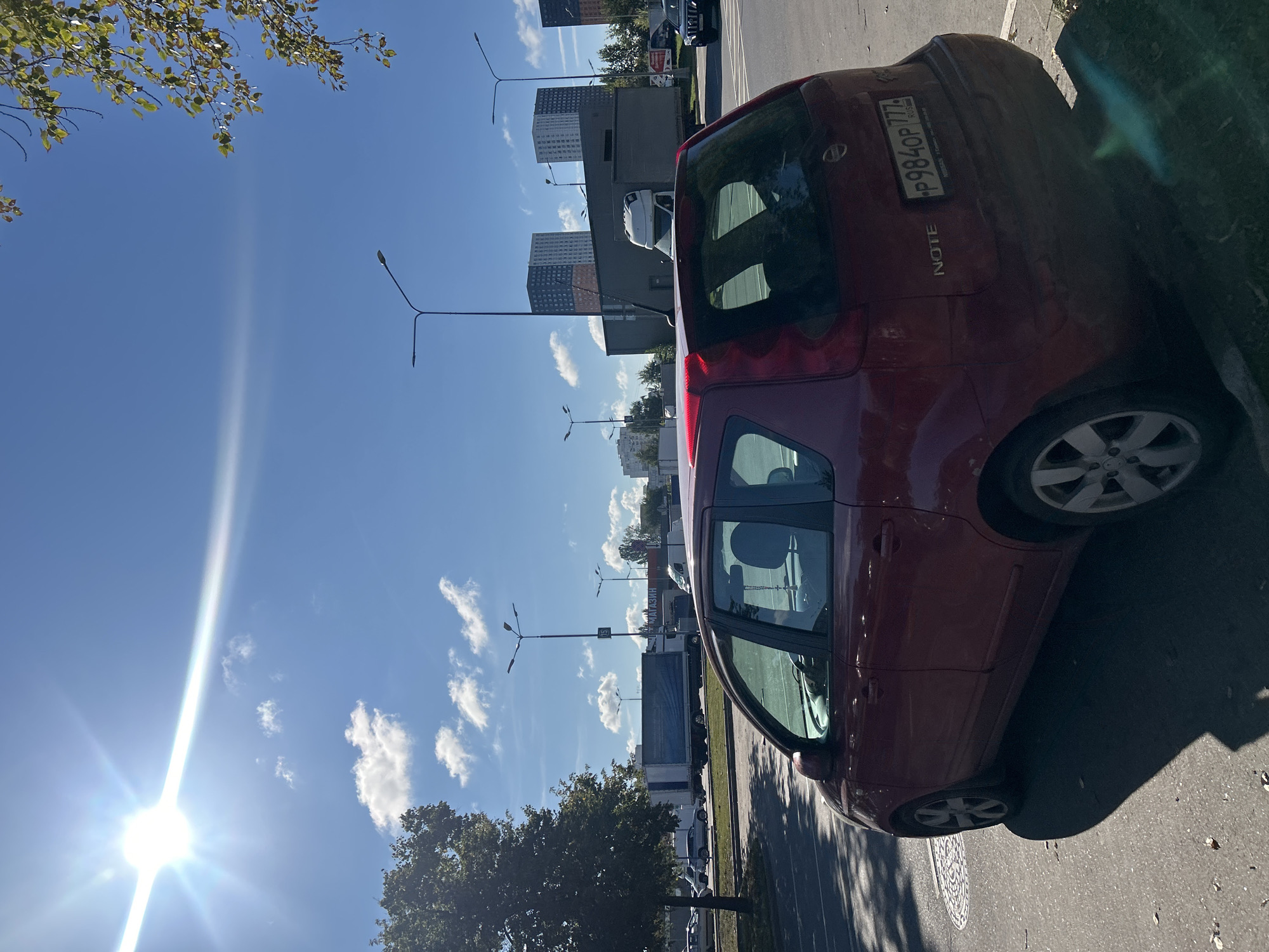This is a medium-shot, eye-level, outdoor, daylight photograph taken on a clear, sunny day. The scene depicts the rear of a red Nissan Note parked on a paved surface, possibly a street or parking lot. The car is positioned at an angle, with its rear facing towards the right side of the frame. A license plate with the characters "P984OP 777 RUS" is visible on the rear bumper.
To the left of the car, a large tree with green foliage casts a shadow. The ground is asphalt with scattered dry leaves and a circular manhole cover visible in the foreground. In the background, a multi-lane road is present, with several vehicles including trucks and cars. Further in the distance, modern residential buildings with distinct orange and blue color schemes are visible, along with streetlights. The sky is a vibrant blue with scattered white clouds and a bright sun that creates a lens flare across the upper portion of the image. The overall setting appears to be a suburban or commercial area of Moscow, Russia. There are no people or animals visible in the image.