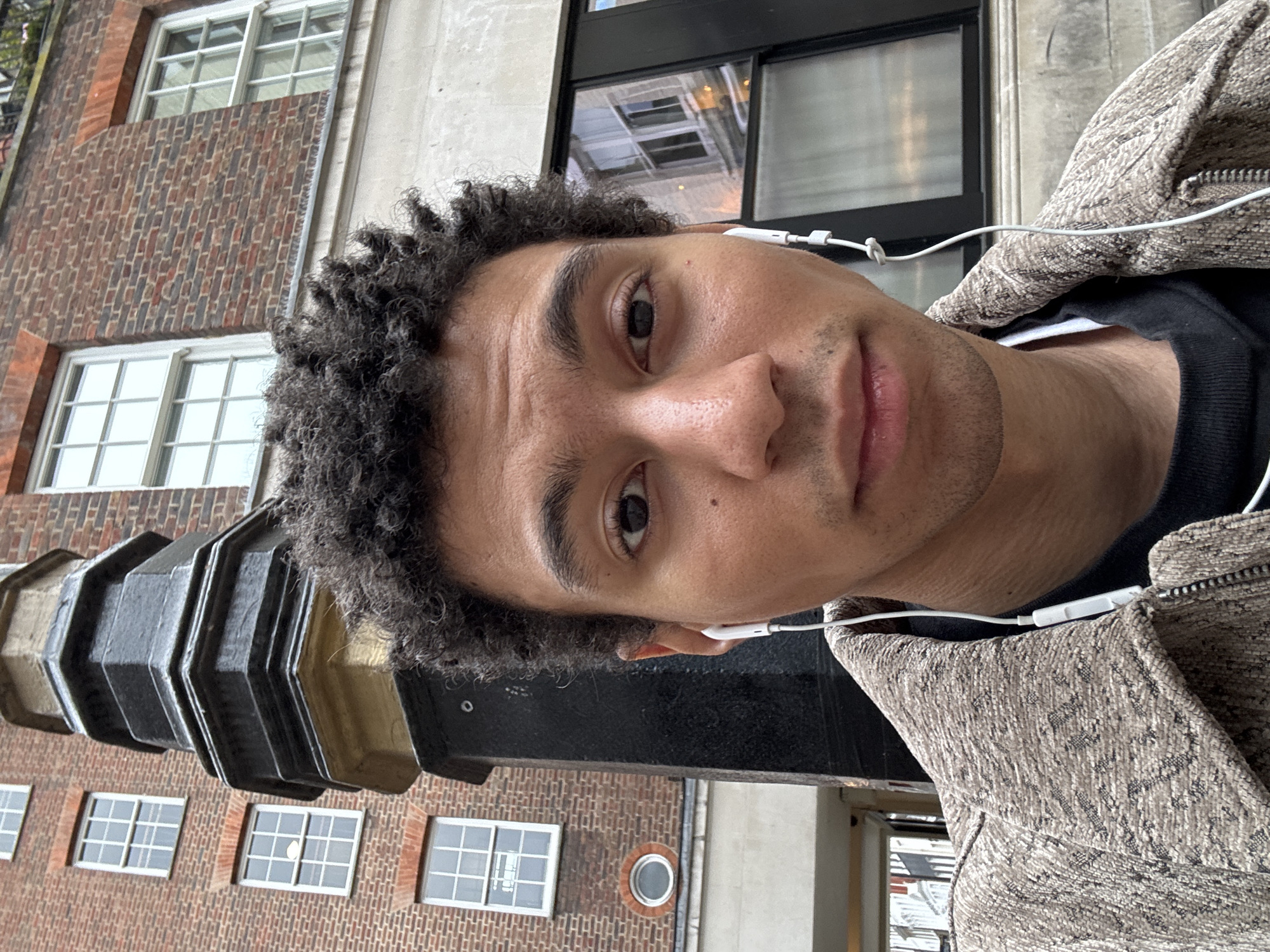 A man is looking at the camera, wearing earbuds and a zippered jacket. He is positioned outside a brick building with multiple windows. The building appears to be a traditional architectural style, with black metallic accents and a small round window. The location appears to be London, United Kingdom.