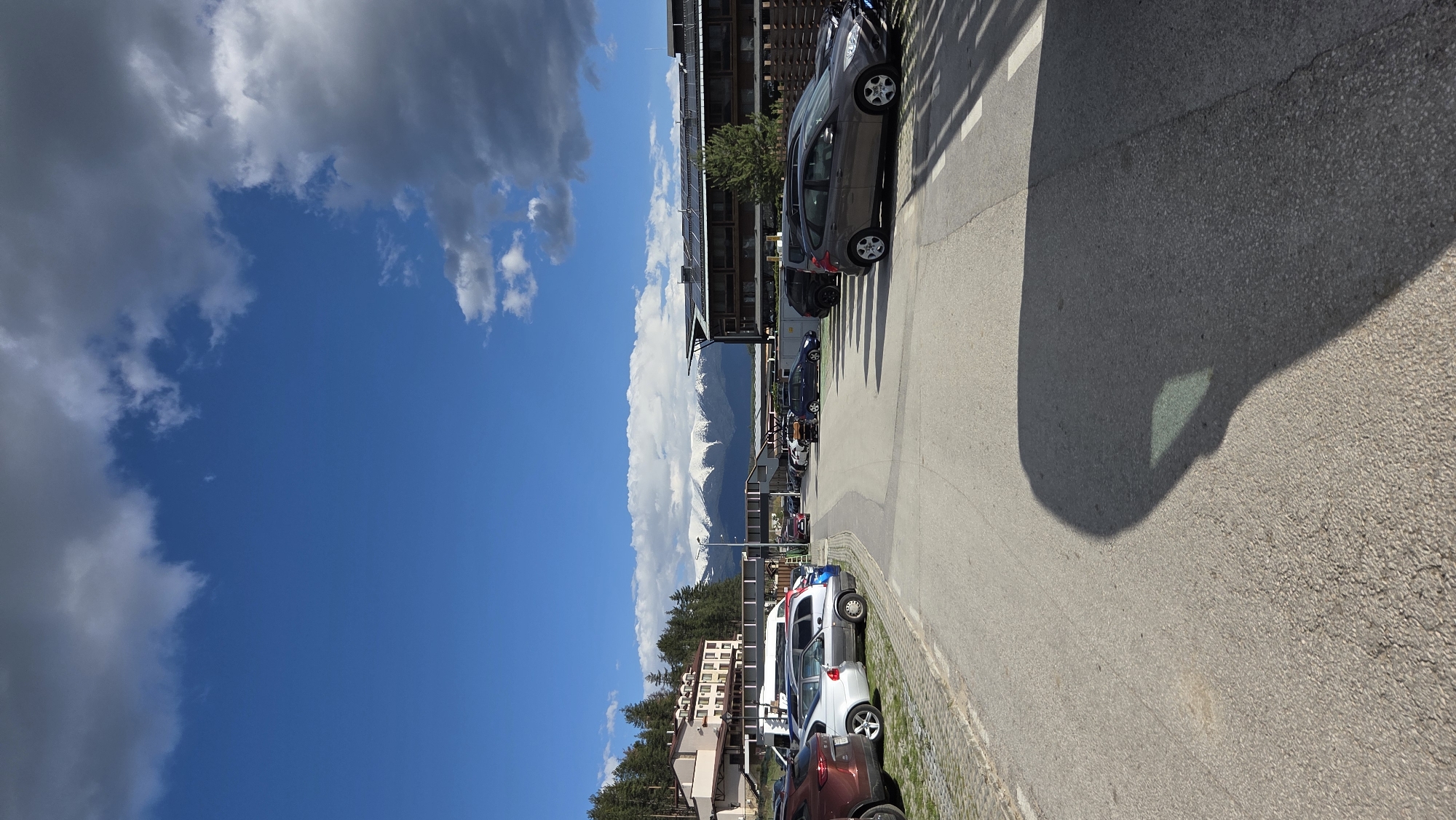 The image captures an outdoor scene in an urban or suburban setting. The most prominent feature is the presence of numerous parked cars lining the side of a paved road. The vehicles are of various makes, models, and colors. In the background, buildings and residential structures are visible, suggesting an inhabited area. The sky is bright blue with patches of white clouds, indicating fair weather. In the distance, there are snow-capped mountains. A large shadow is cast onto the road, obstructing some of the view.
