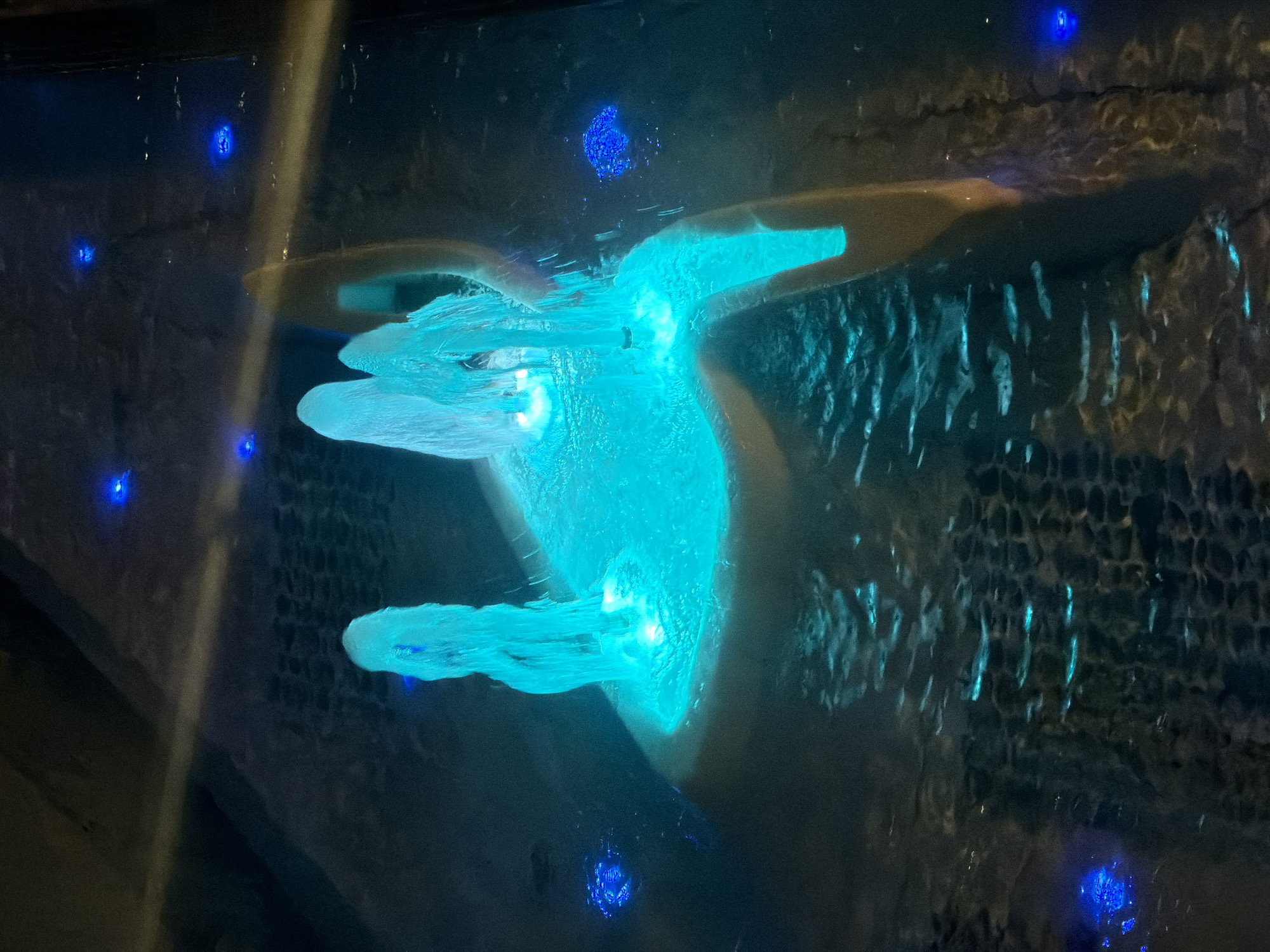 The image shows an illuminated water feature at night. Multiple jets of turquoise water shoot upwards and outwards, creating a dynamic and fluid sculpture. The water is lit from below by bright, glowing turquoise lights, which also illuminate the surrounding pool and a light brown, curved structure. Several small, bright blue lights are scattered across the surface of the water and in the background. The pool floor is covered with dark, textured material, possibly stones or tiles, which are also partially illuminated. A diagonal beam of yellowish light cuts across the scene from the upper left. The overall impression is one of a modern, artistic water installation designed for visual appeal in a dark setting. Based on visual cues, this appears to be the fountain at Eleftheria Square in Nicosia, Cyprus.