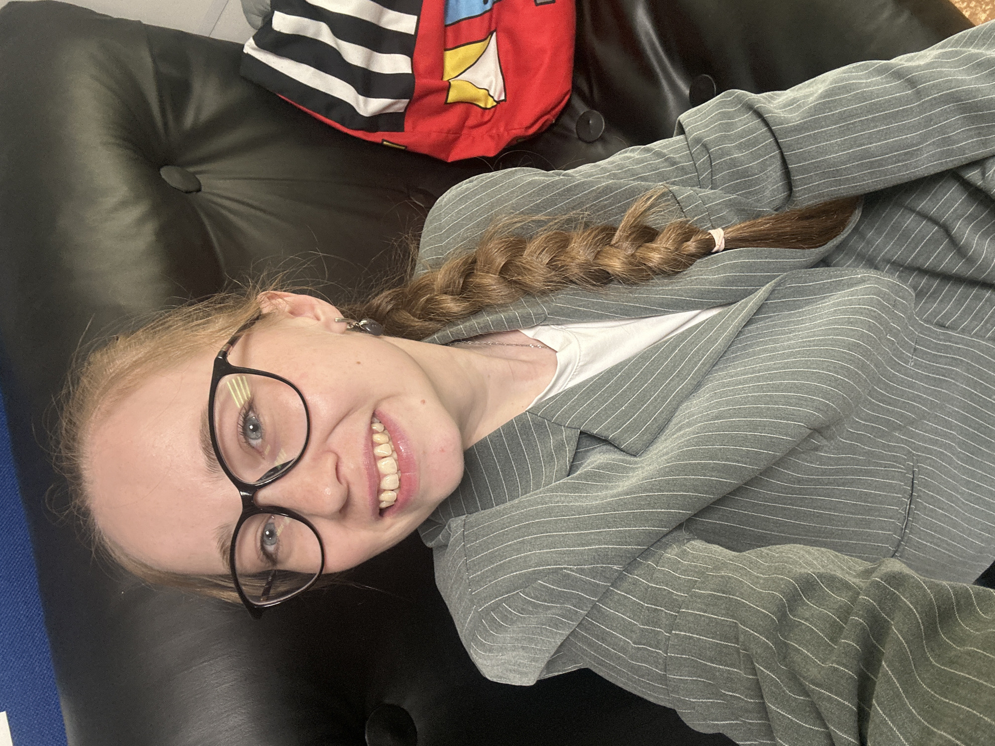 The image shows a young woman with fair skin, light brown hair styled in a braid, wearing black-rimmed glasses, a white t-shirt, and a gray pinstripe blazer. She is smiling and appears to be lying on a black, button-tufted surface. To the left of her head, there is a glimpse of blue material. Behind her, there is a red and black striped object. The image does not provide enough information to determine if this scene is in Saint Petersburg, Russia.