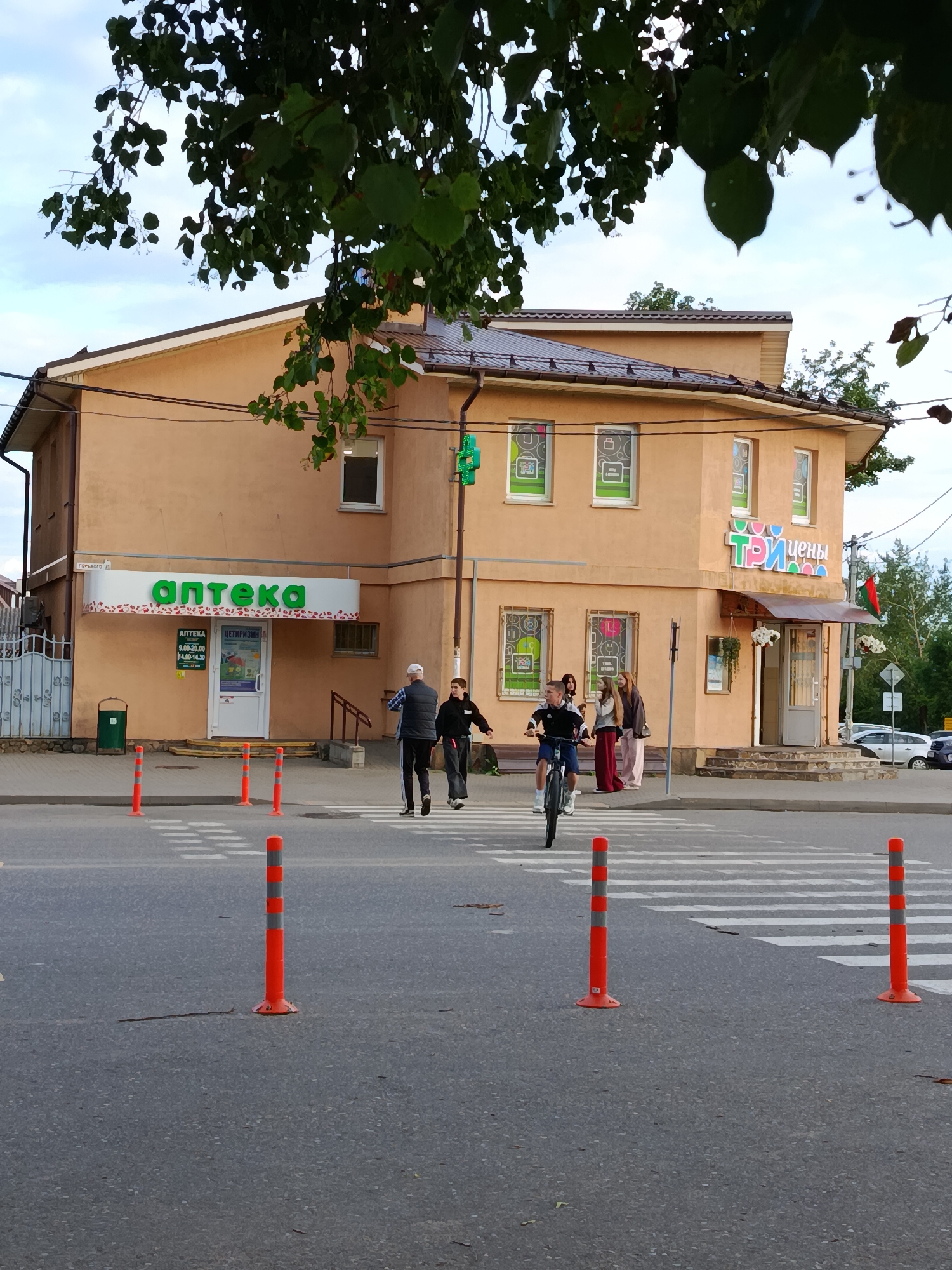 An exterior, eye-level, medium shot captures a street scene in front of a two-story commercial building. The building, painted a peach color, has a dark roof and multiple windows. A prominent green sign reading "аптека" (pharmacy) is visible on the left side of the building. On the right side, a colorful sign displays "ТРИ ЦЕНЫ" (three prices). Several people are present: a man in a blue jacket and black pants, a boy in a black hoodie, a girl in a white hoodie and pink pants, and another girl in a brown hoodie and maroon pants. A young man is riding a bicycle across a pedestrian crossing in the foreground. Orange and gray striped traffic cones are placed on the road, delineating the pedestrian crossing. The sky is cloudy. A lush green tree hangs over the top of the building, with its leaves obscuring a portion of the sky. The location is the city of Смілавіцкі сельскі Савет, Belarus.