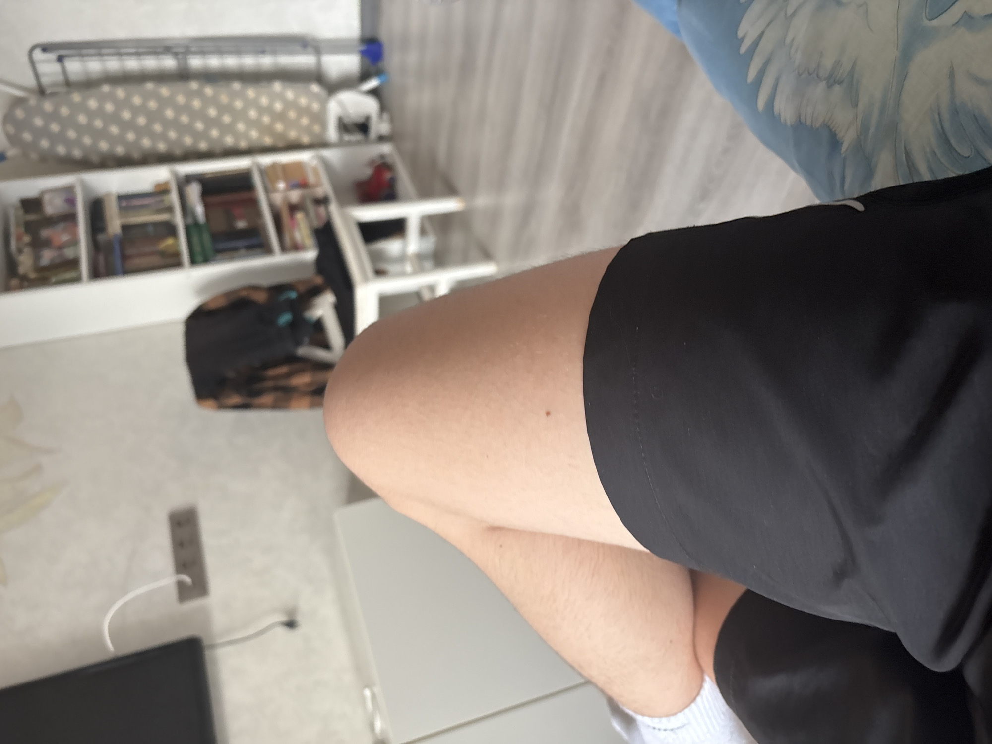 The image shows a person's legs, clad in black shorts and white socks, resting on a blue and white patterned surface. The legs are positioned with the right leg crossed over the left. In the background, a white bookshelf filled with books and decorative items stands against a light-colored wall. To the left of the bookshelf, a white stool with a black cushion is visible. Further to the left, a white cabinet or dresser is present, with a wall-mounted electrical outlet and cable on the wall above it. The flooring appears to be light-colored wood planks. The overall setting suggests an indoor residential environment.