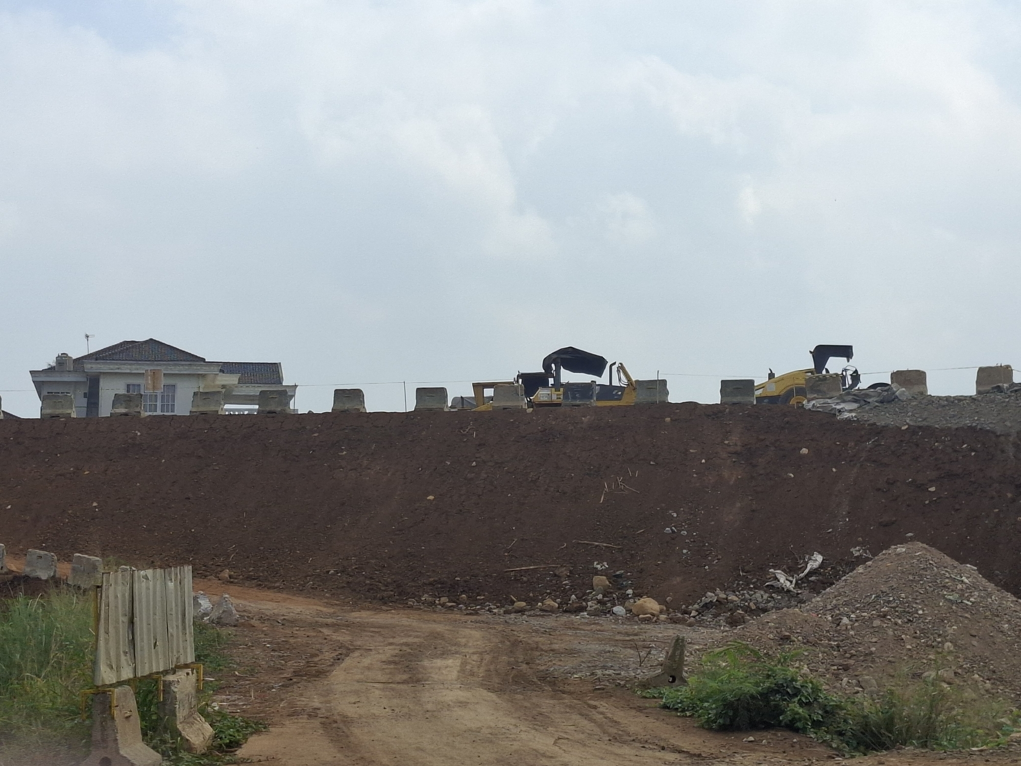 The image depicts a construction site in Ciheulang Tonggoh, Indonesia, characterized by a large mound of dark brown soil, likely for earthworks. Several heavy construction vehicles, including yellow excavators and bulldozers, are visible on top of the mound, some partially covered by black tarps. In the background, a white building with a dark roof, possibly a house or office, is situated behind a barrier of concrete blocks along the edge of the construction area. The sky above is overcast with clouds. In the foreground, a dirt path leads towards the mound, with some overgrown vegetation and more concrete barriers.