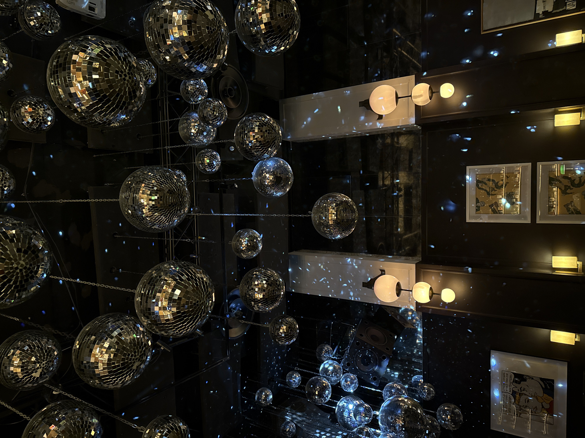 The image shows a room filled with disco balls, creating a mirrored and reflective environment. The lighting fixtures on the walls are notable, and framed artwork adorns the surfaces. The space seems to be designed for entertainment or a nightclub-like atmosphere.
The text partially visible in a framed artwork reads, "МИЛЫЙДА СПАСИБО, КРОШИ ПАРОСТРУ ПЕНИЙ НО ВСЁ-ТАКИ Я НЕ ЭТО ШЕДЕВРА ЛИХТЕНШТЕЙН/", which is Russian for "Dear, thank you, crumbs of steam singing, but I don't This is a Lichtenstein masterpiece".
Based on the text in the artwork being in Russian, the location is likely in Moscow, Russia.