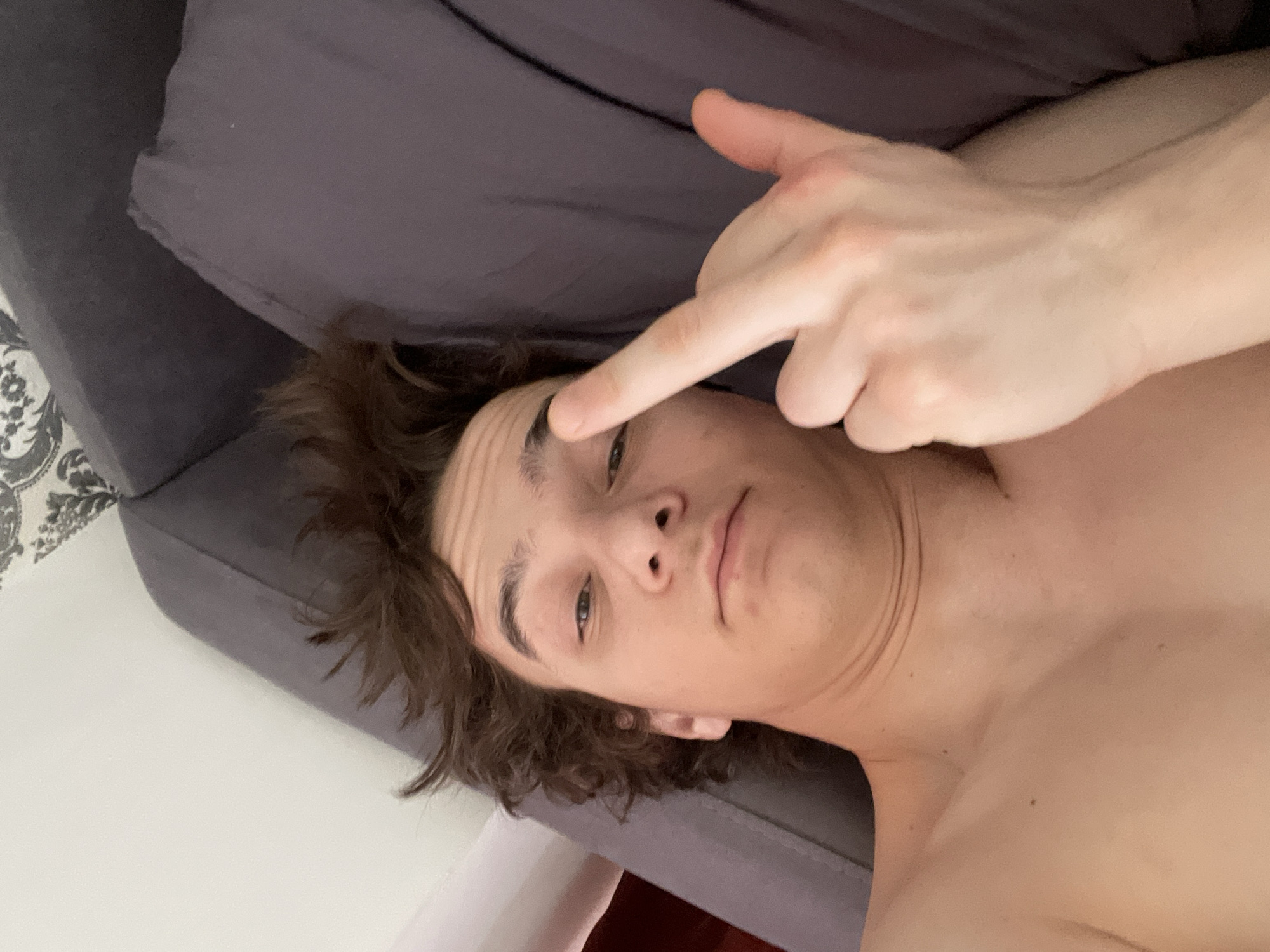 An indoor, overhead shot captures a young individual lying horizontally, likely on a bed or couch, in what appears to be Krasnoyarsk, Russia. The person, who has brown, somewhat curly hair, is shirtless, revealing their bare chest and shoulders. Their left hand is raised, with the index finger pointing towards their right eyebrow, while their expression is neutral and contemplative.
The immediate background consists of a dark grey or purplish fabric, likely a pillow or bedding, and the frame of a bed or sofa. To the left, a white wall or sheet is visible, along with a fragment of a white and grey patterned rug or upholstery. The lighting is soft and diffused, creating subtle shadows and highlighting the person's features, including their prominent eyebrows and visible forehead lines. The overall impression is candid and relaxed, capturing a personal moment. No text is visible in the image.