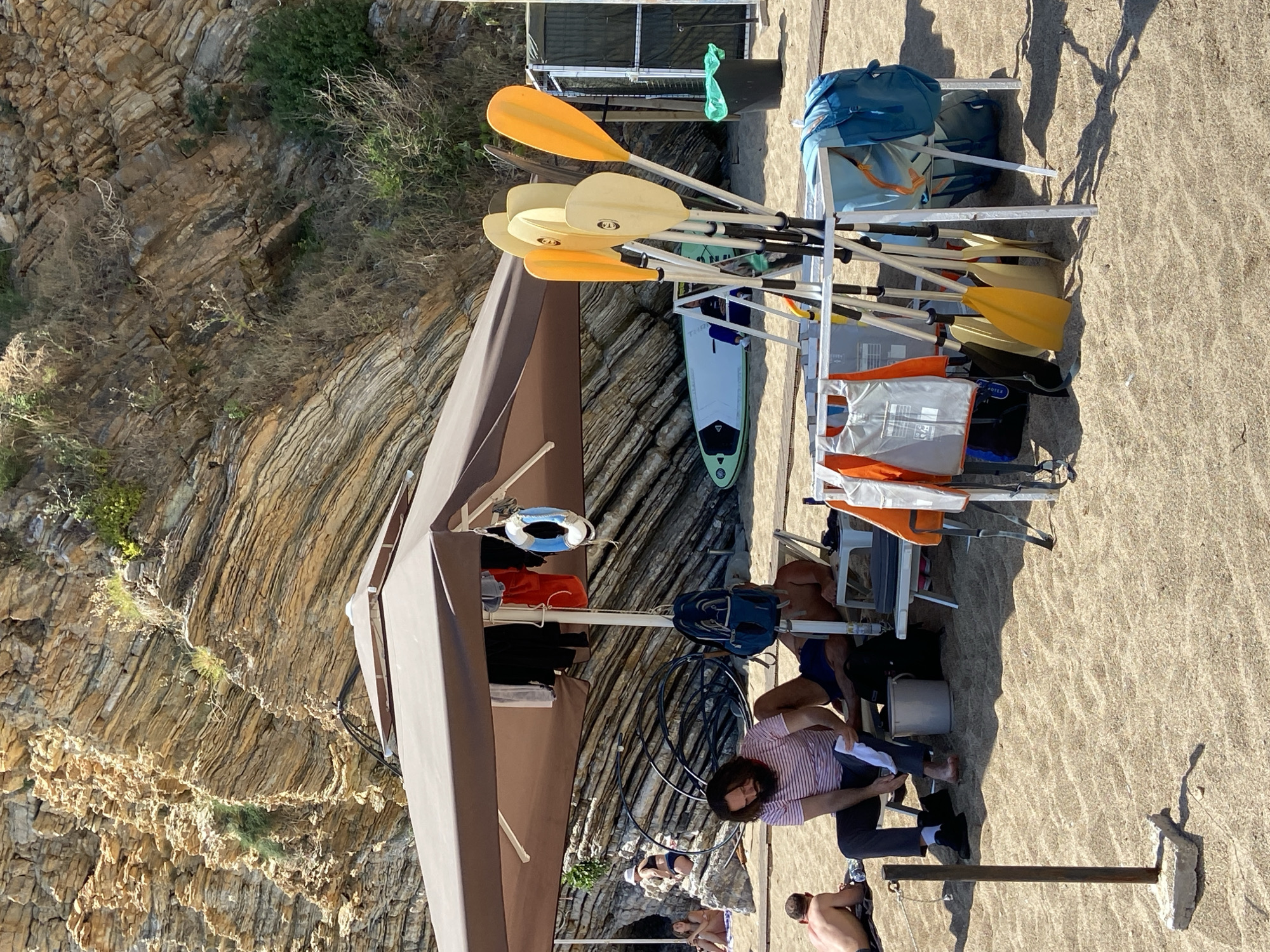 The scene appears to be on or near a beach in Budva, Montenegro, indicated by the coastal rock formations and sandy ground. Several people are present, some seated near a building with a brown awning. There's a metal storage rack laden with items: paddles of different colors, possibly for kayaks or paddleboards; blue bags; and orange life vests. A surfboard is also visible. Other beachgoers appear to be sunbathing in the background, partially obscured by the awning. A man sits on a chair near the rack.