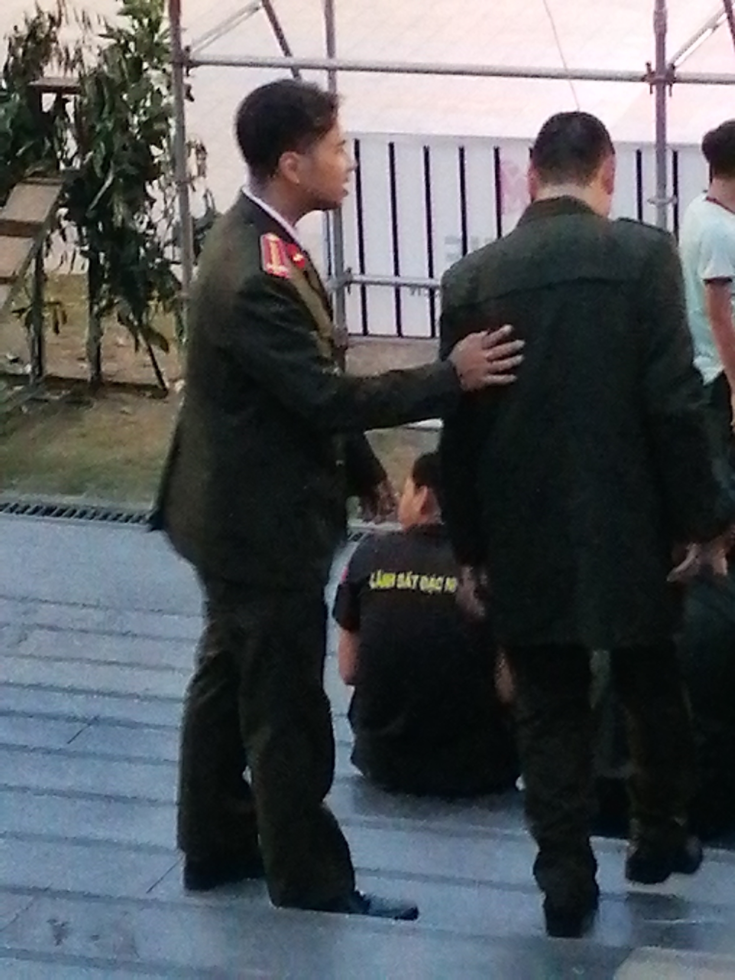 Two men in dark green uniforms with red and gold epaulets are the primary subjects in this medium shot. The man on the left, facing the right, has his hand placed on the shoulder of the man on the right. The man on the right is facing away from the camera, with his arm around the man on the left. Both men appear to be wearing dark pants and shoes. Behind them, another person in a dark t-shirt is seated, with visible text on the back of the shirt. The setting appears to be outdoors, with a backdrop of a white tent or structure with black vertical bars, and some greenery to the left. The lighting suggests it might be daytime. The overall scene is somewhat blurry, with a focus on the two uniformed men. The visible text on the t-shirt is in Vietnamese.