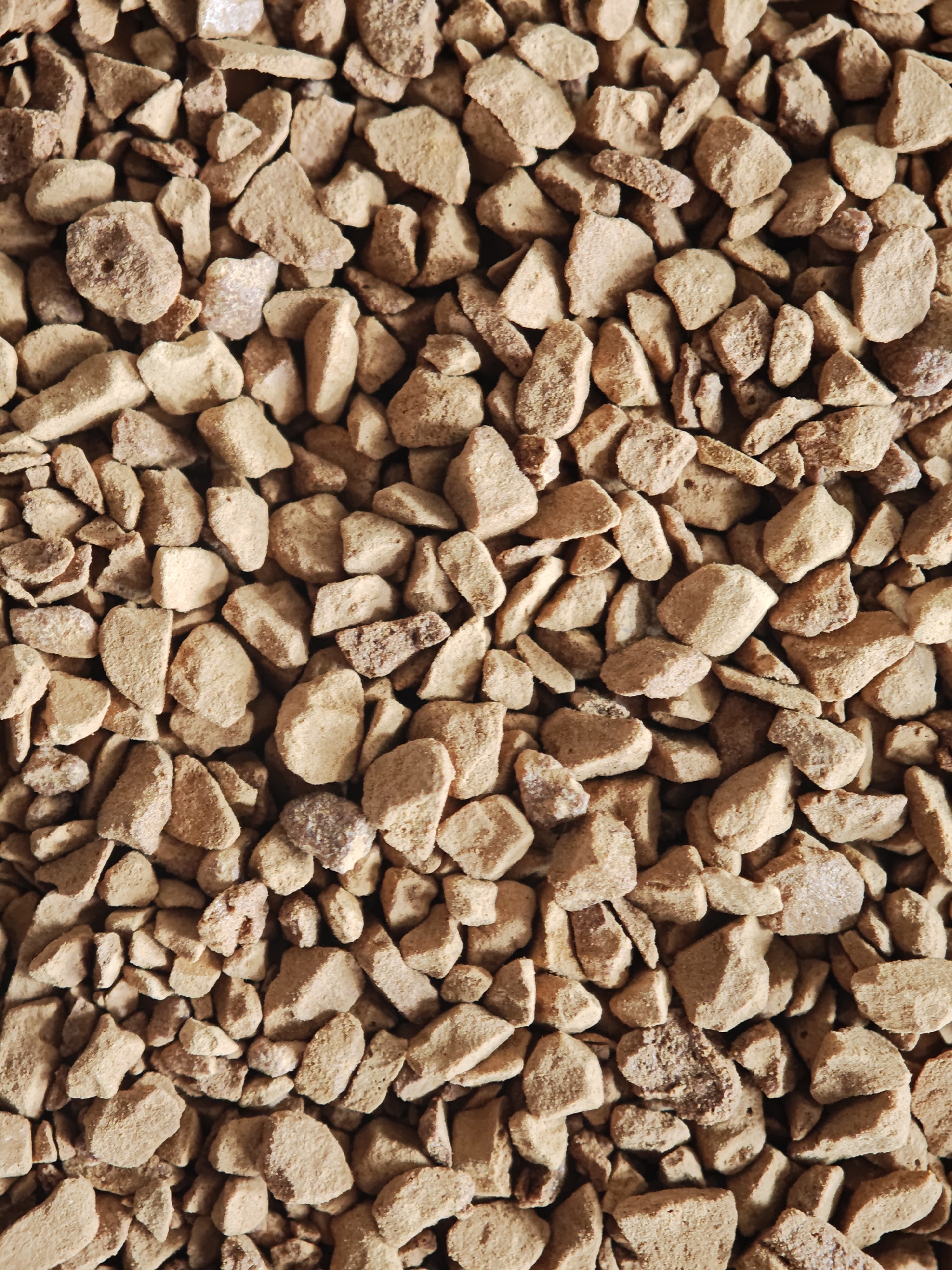 The image is a close-up, overhead view of a textured surface composed of numerous small, irregular, tan-colored granules. These granules appear to be coarse and somewhat porous, with varying shapes and sizes, suggestive of instant coffee granules or similar food product. The lighting creates subtle shadows, highlighting the three-dimensional nature of the material. There is no discernible background or environmental context visible. The image does not depict any location in the city of Kursk, Russia, nor does it contain any people, actions, or interactions.