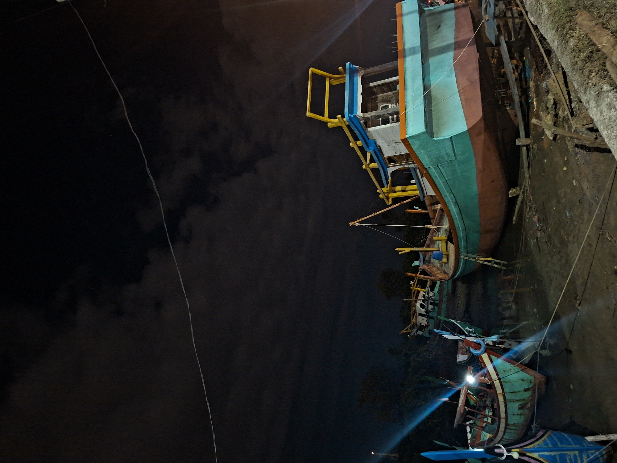The photograph depicts a night scene in Lhokseumawe, Indonesia, featuring a grouping of brightly painted fishing boats on the shore. The largest vessel, in the upper right, is elevated on a wooden frame and displays teal, brown, and orange colors, with yellow and blue structures. Lower down, another boat shines brightly with teal and red trim under spotlight illumination. The backdrop consists of a darkened sky with indistinct clouds, and a powerline bisects the sky to the left of the boats. The ground is dark and uneven. The scene is mostly focused on boat construction.
