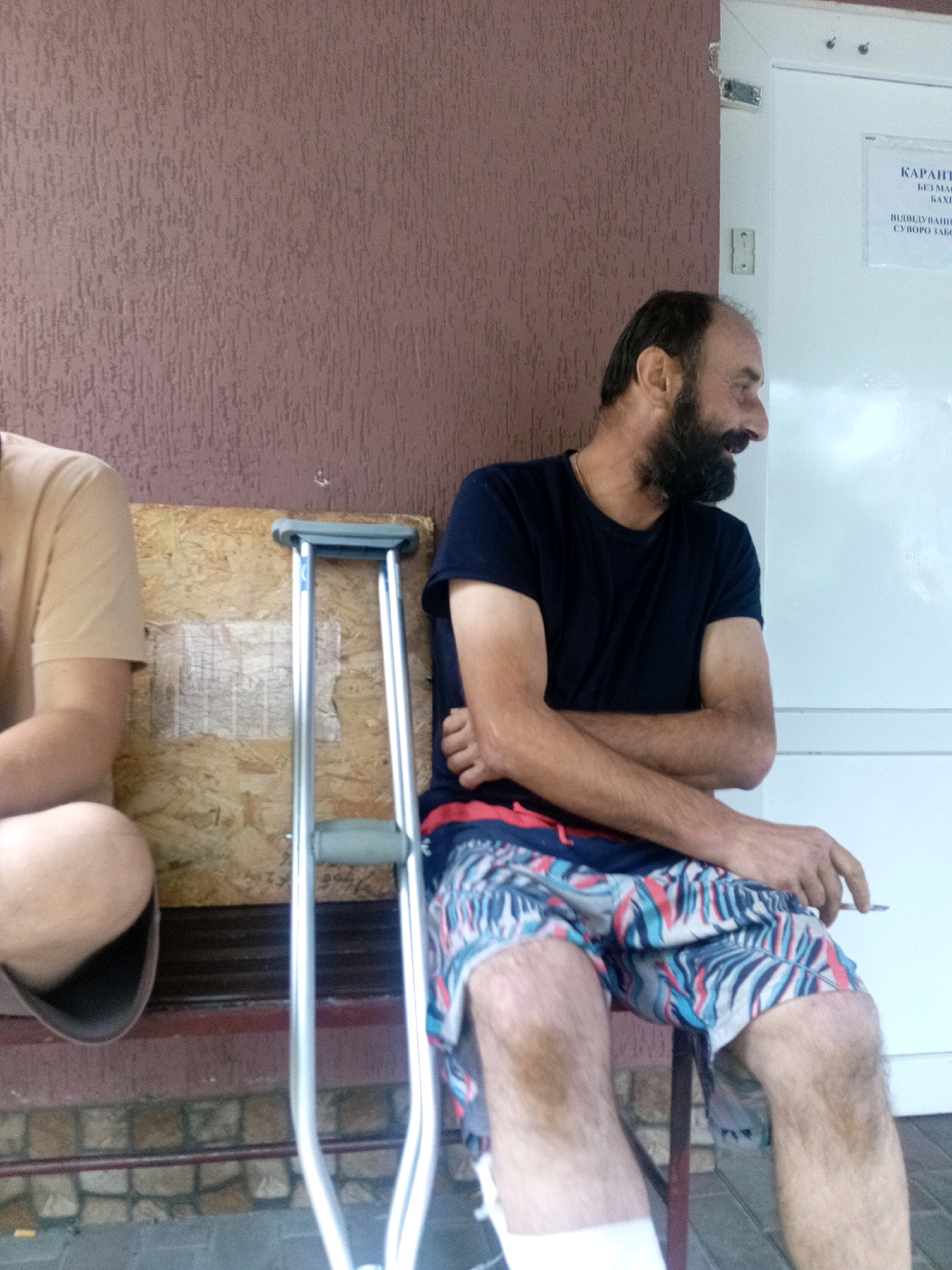 Two individuals are seated outdoors on a low wall or bench, with one person in the foreground. The person in the foreground is a man with a beard, wearing a dark blue t-shirt and patterned shorts. He is holding a cigarette in his right hand and appears to be smoking. His arms are crossed. To his left, a portion of another person is visible, wearing a light brown t-shirt. Between the two individuals is a silver-colored crutch. Behind them is a textured brown wall and to the right, a white door with a sign that reads "КАРАНТИН" (Quarantine) and some additional text in Ukrainian. The ground in front of the bench appears to be paved with stones. The overall scene suggests a moment of rest or waiting in an outdoor setting. The location is identified as Tiachiv, Ukraine.