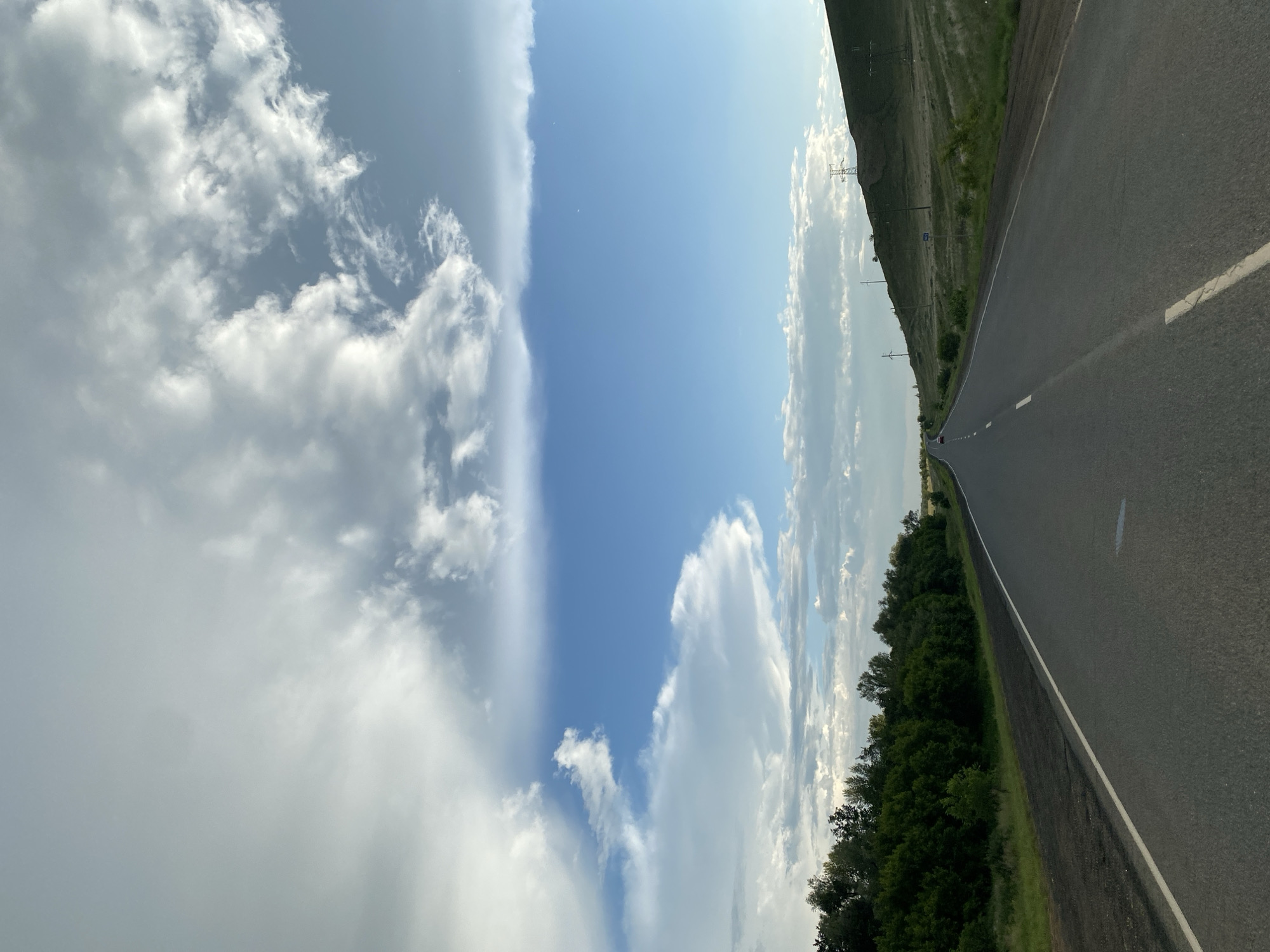 This is a forward-facing, eye-level shot of a paved road leading into the distance. The road has a white dividing line and a white line along the right shoulder. To the left of the road is a dense line of green trees. To the right, the land slopes upwards into a grassy hill with several power lines visible. The sky is a bright blue with large, dramatic white clouds, some appearing wispy and others more dense. The overall impression is a scenic drive in a rural or semi-rural area, possibly on the outskirts of a city given the presence of power lines and a road suitable for travel. The location is stated to be in Verkhneozyornoye, Russia, though there are no specific landmarks to confirm this within the image itself beyond the general landscape. There are no people visible, and the scene is static, depicting a moment of travel.