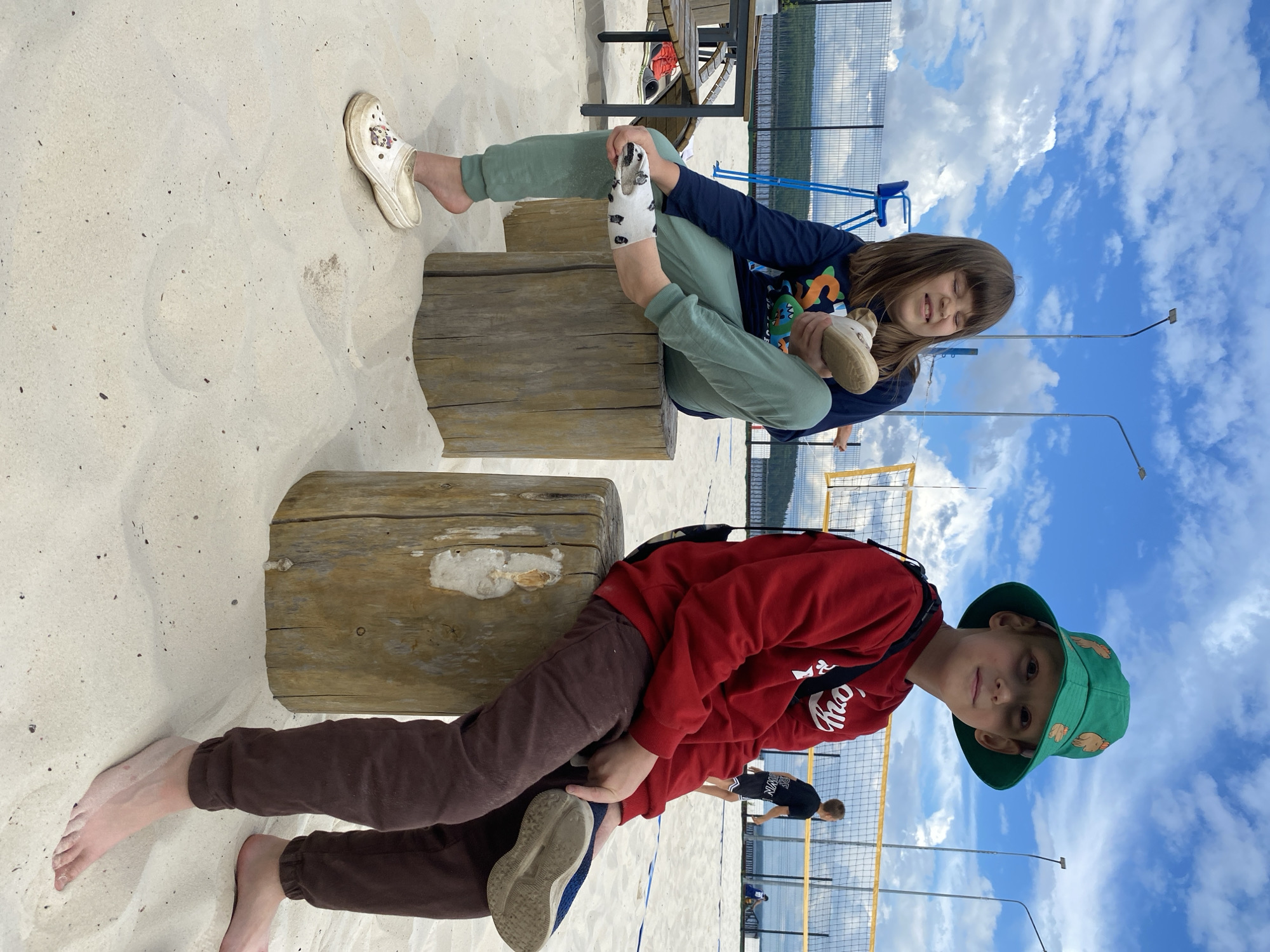 Two children are sitting on logs on a sandy beach with a volleyball net in the background. The child on the left is a girl with long brown hair and is wearing a navy blue long-sleeved shirt and light green sweatpants. She is holding a white shoe in her hand and has a white clog on her left foot. The child on the right is a boy wearing a red sweatshirt, brown sweatpants, and a green bucket hat. He is holding a blue shoe. The sky is cloudy and blue. The location is the beach at the Kama River in Chaikovsky, Russia.