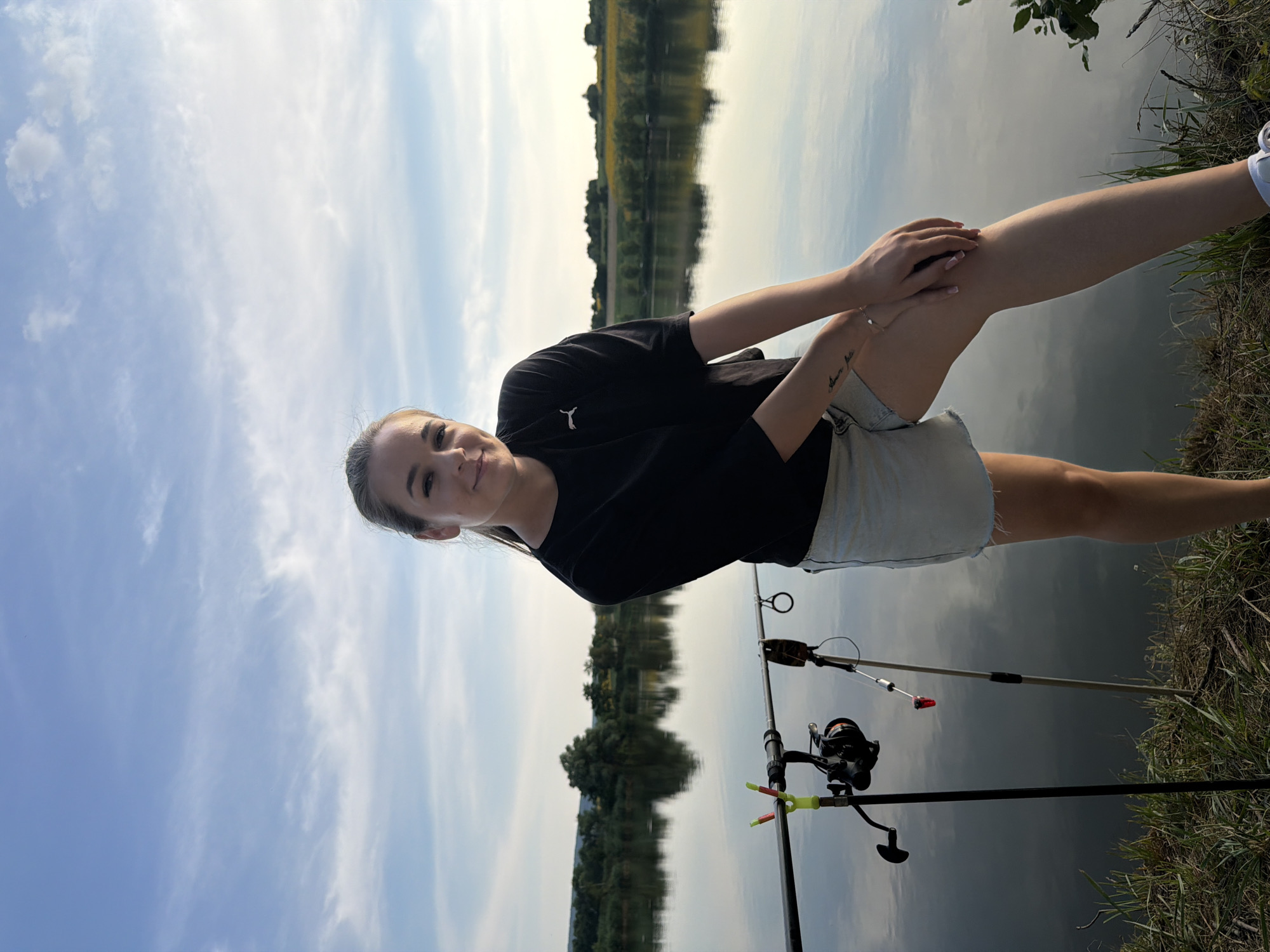 A young woman is standing on the grassy bank of a calm body of water, possibly a lake or river, in Găleștii Noi, Moldova. She is wearing a black t-shirt and light-colored denim shorts. Her hair is pulled back in a ponytail. She is leaning on her right leg, with her left leg bent and resting on her right knee. Her hands are clasped together. There are fishing rods set up on the bank to her left. The water reflects the blue sky with scattered clouds and the green trees and vegetation on the opposite bank. In the background, there is a treeline and fields under a bright sky.