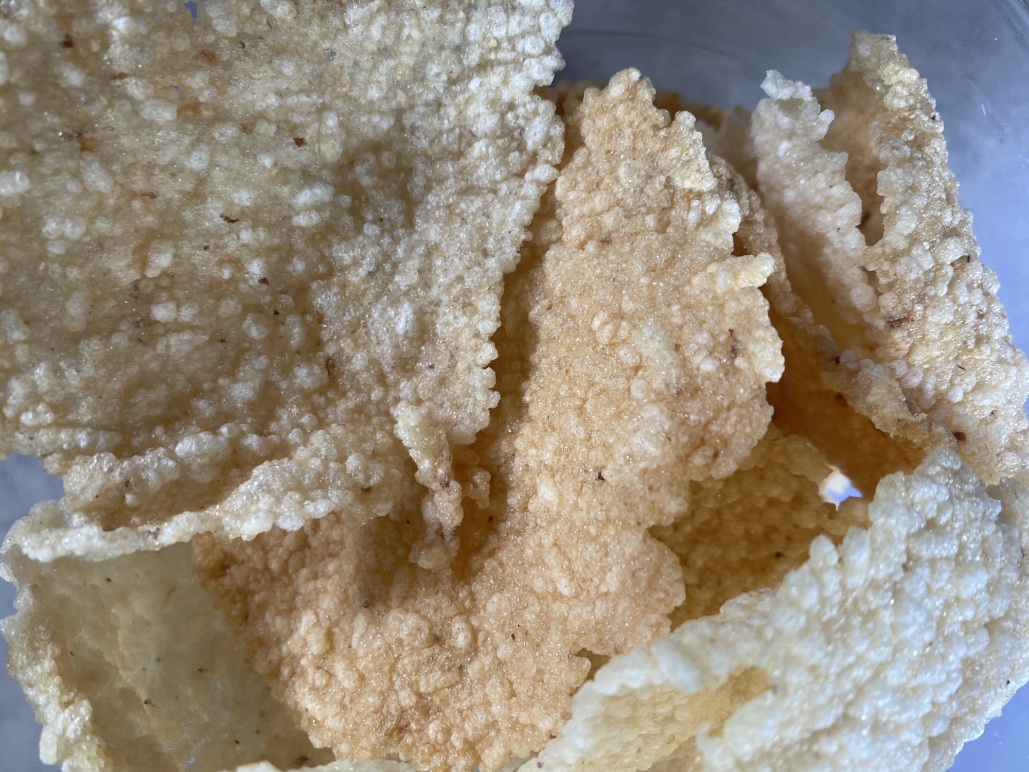 A close-up shot displays a pile of crispy, light-brown and off-white crackers. The crackers have an irregular, porous texture with small, raised bumps across their surfaces. Some of the crackers are layered on top of each other, creating a sense of depth. The lighting suggests an indoor setting. The scene depicts food items, specifically crackers, and does not show any elements related to the city of Kasihan, Indonesia.