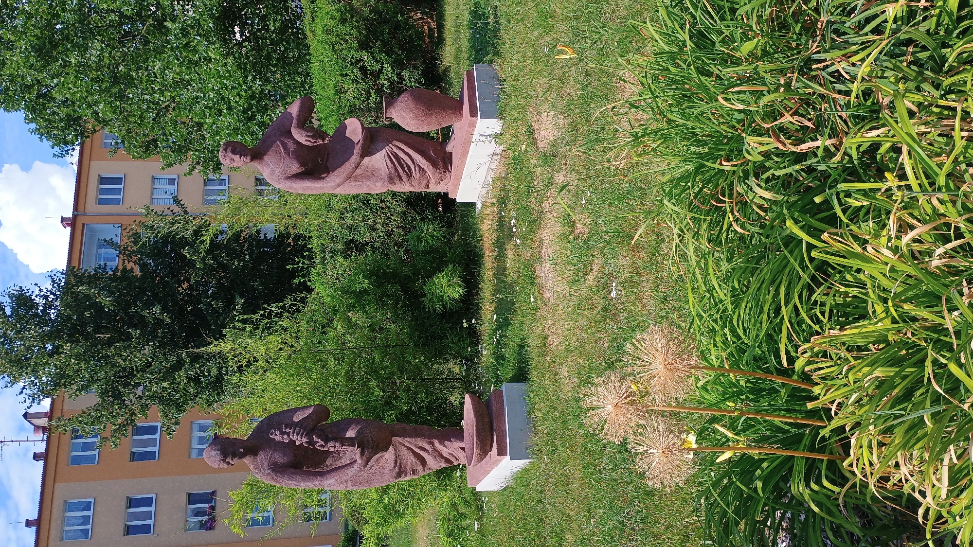The image shows two reddish-brown stone statues of women in a grassy area in front of a light yellow and orange apartment building. The statue on the left depicts a woman holding a bundle of what appears to be reeds or grain. She stands on a white pedestal with a pinkish-red top that has a shallow bowl on it. The statue on the right shows a woman holding a shallow bowl in one hand and reaching towards a large vase with the other. She also stands on a white pedestal with a pinkish-red top. Behind the statues are dense green bushes. In the foreground, there are ornamental dried grass seed heads and lush green foliage. The sky is blue with some white clouds. The building appears to be a multi-story residential structure typical of Eastern European cities. The overall scene suggests a public park or communal garden area. Based on visual cues, this scene is located in the city of Sokolov, Czechia, likely in a residential district.