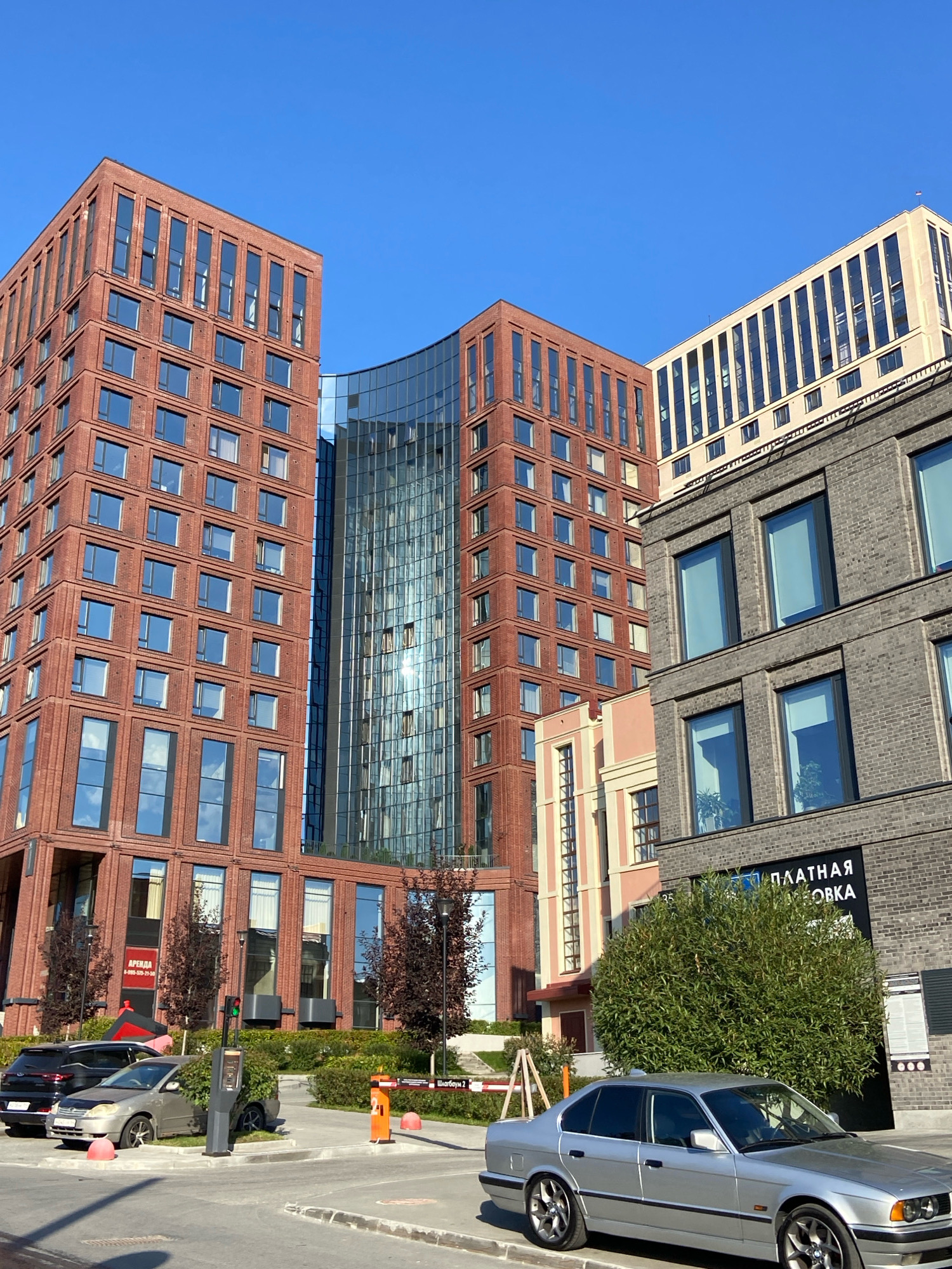The image depicts a modern urban scene with several multi-story buildings under a clear blue sky. On the left, a prominent building is constructed with red brick, featuring numerous rectangular windows with dark frames. Adjacent to it, a curved section of a building showcases a glass facade that reflects the sky and surrounding structures. To the right, another building is made of dark grey brick, also with large, rectangular windows. A lighter colored building with vertical windows is visible further in the background.
In the foreground, a silver sedan is parked on a paved surface. There is a parking barrier with an orange pole and red bollards. A traffic light with red and green signals is visible near a control box. Several trees with dark foliage are planted around the buildings. A sign with Cyrillic text, partially obscured by a tree, is visible on the grey brick building. The overall environment suggests an organized, possibly commercial or residential complex in a city.