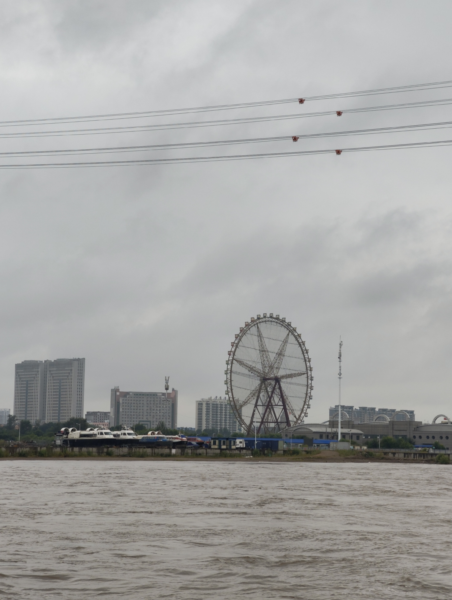 The image depicts a riverside view with a city skyline under a cloudy sky. In the foreground, a body of murky, choppy water dominates the lower half of the frame. Along the riverbank, a row of hovercrafts are parked, with several tall buildings visible behind them. To the right of the buildings, a large Ferris wheel stands prominently. Above the cityscape, several sets of power lines stretch across the frame, with small red insulators attached to them. The environment appears to be industrial or a mixed-use urban area located in 幸福乡, China, given the presence of the Ferris wheel and the urban development along the river. The weather is overcast, casting a muted light on the scene. There are no people or animals visible in the image.