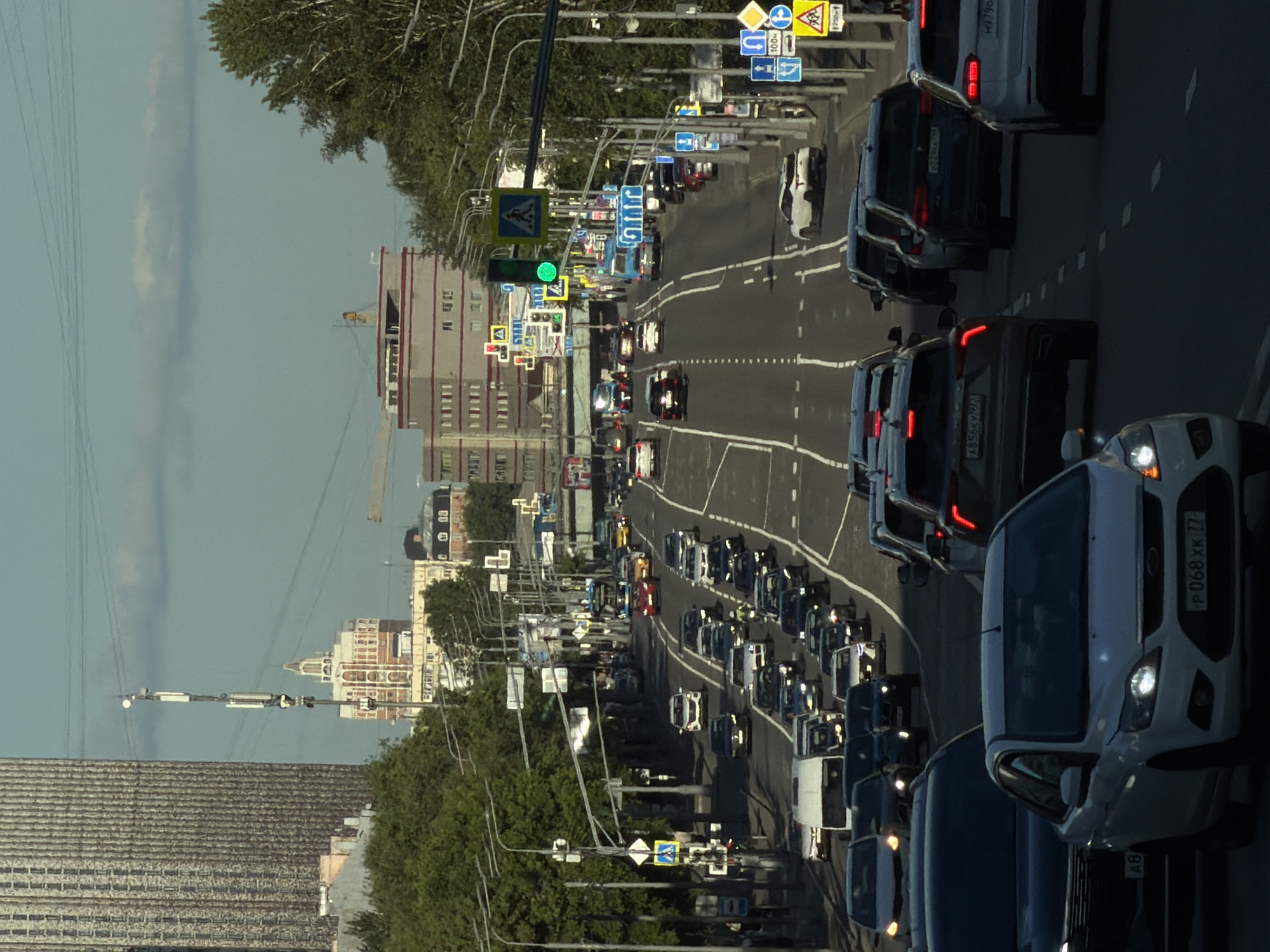 This is a medium shot, taken from an elevated position looking down at a street in Khimki, Russia. The scene depicts a busy roadway with numerous cars moving in both directions, suggesting a rush hour or significant traffic. The orientation of the image is such that the cars are traveling vertically from the bottom to the top of the frame, and vice versa.
On the right side of the road, several cars are visible, including a white van or SUV with its headlights on. Further down the road, more vehicles are lined up, some with their brake lights illuminated. On the left side of the road, a row of trees lines the street, with a modern building visible behind them. Further in the background, taller buildings with distinct architectural features can be seen, characteristic of urban Russian architecture. A network of overhead wires or power lines also crisscrosses the upper part of the image.
Traffic signs are prominently displayed along the road, including directional signs and road safety symbols. A traffic light is visible, showing a green signal. The sky appears clear and bright, indicating it is daytime, likely late afternoon or early evening given the angle of the sunlight casting shadows. The overall atmosphere is one of urban activity and movement.
Visible text on the license plate of the white vehicle in the foreground reads "P 068 XK 77". Other license plates are partially visible on other vehicles, but the text is illegible.