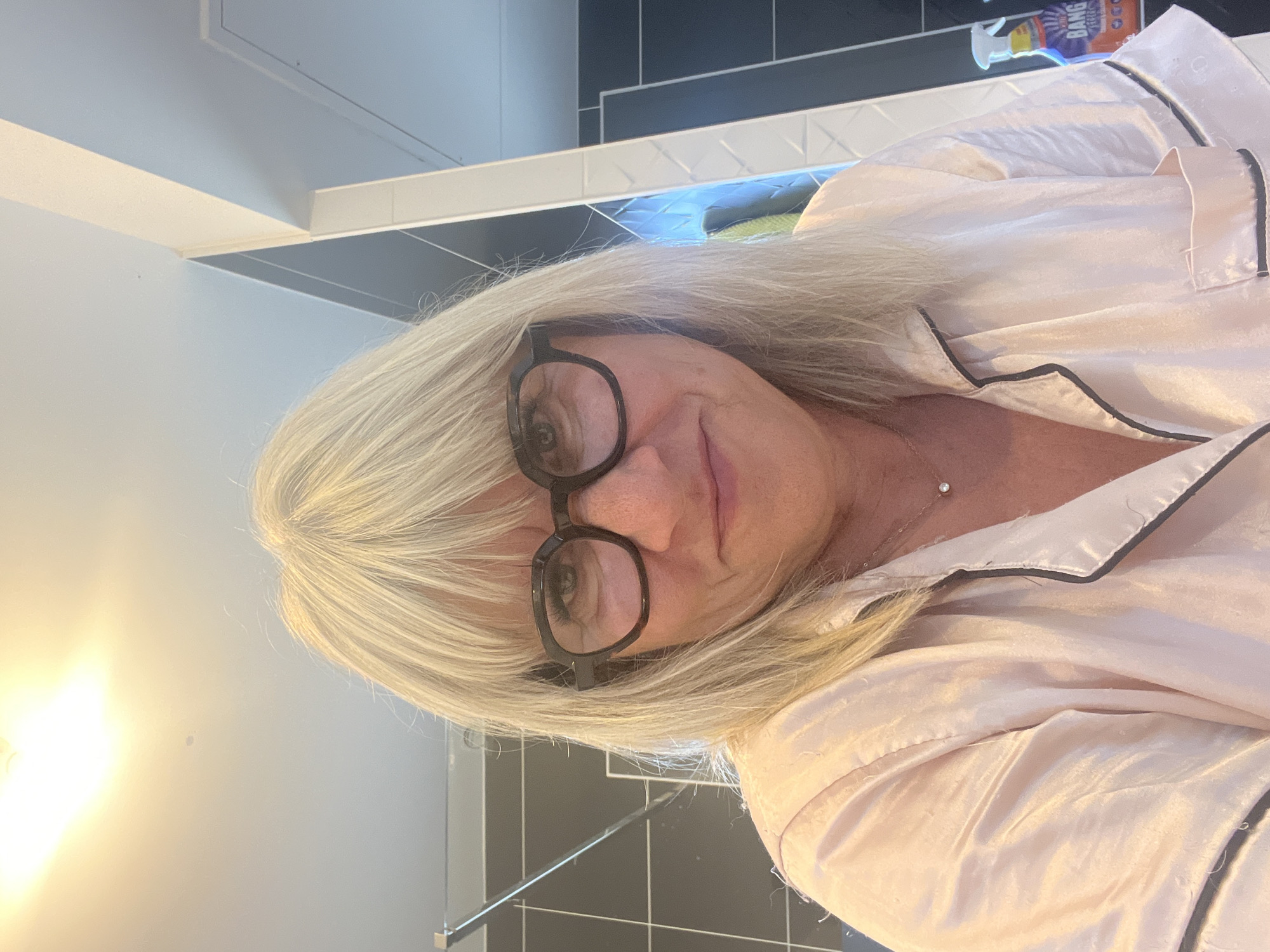The image shows a close-up, selfie-style shot of a woman with blonde hair and glasses. She is wearing a pink satin pajama top with black trim. She has on a delicate necklace with a small pendant. The background appears to be a bathroom with tiled walls and a bright overhead light. There is a bottle of cleaning spray visible on the right side of the image. The location is not specified as Chambœuf, France; this detail is extraneous and cannot be determined from the image itself.