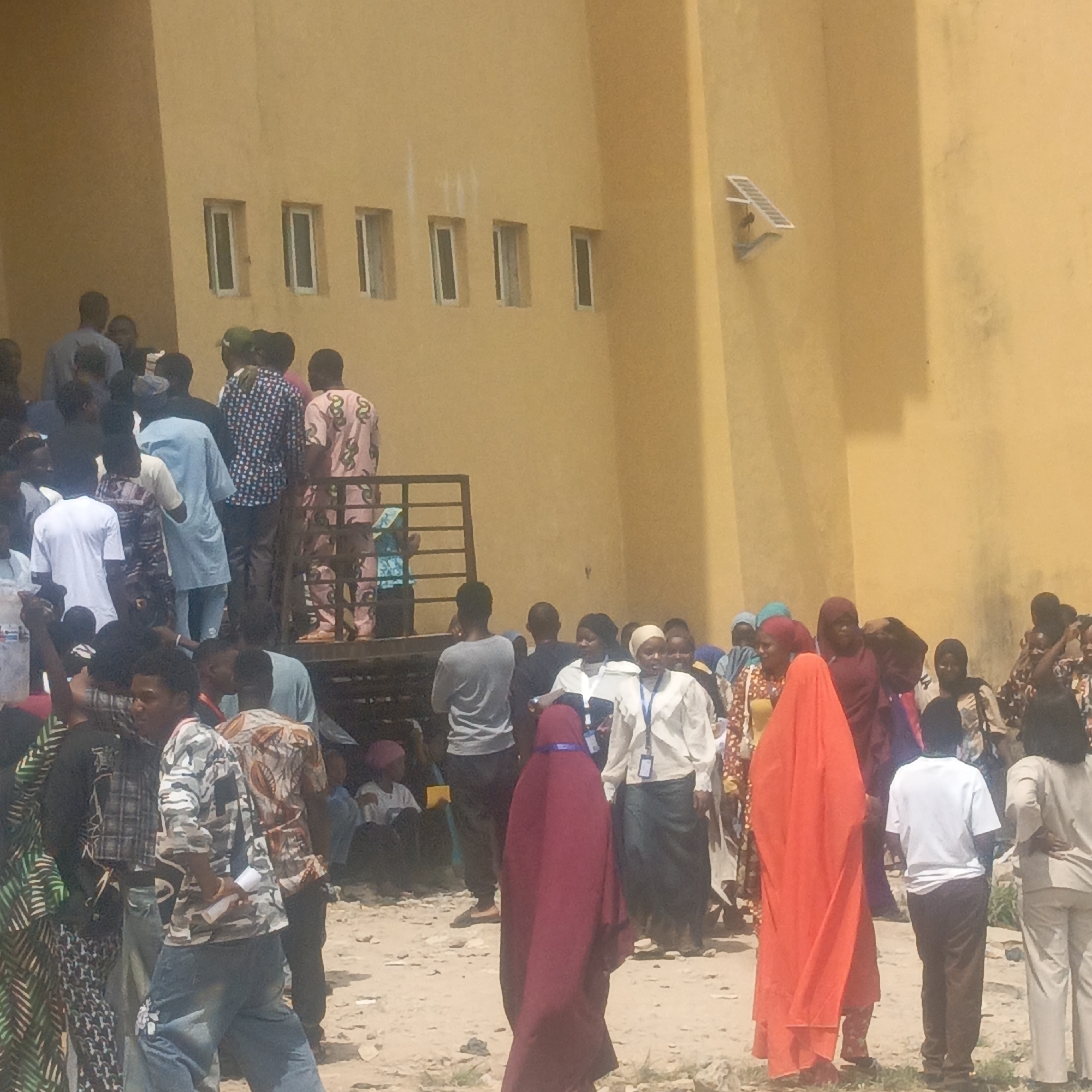 The image captures a bustling outdoor scene in Keffi, Nigeria, under bright daylight. A large group of people, appearing to be mostly adults, are gathered in front of and around a plain, yellow-painted building. The ground beneath them is sandy or dusty.
Many individuals are standing, suggesting they are waiting or queuing for an event or service. On the left side of the frame, several people are seen ascending or standing on a metal staircase or platform attached to the building. The attire of the crowd is diverse; some women wear traditional head coverings like hijabs, and two women in the foreground are notable for their full maroon and orange burkas/niqabs. Several individuals, particularly in the mid-ground, wear lanyards with ID badges around their necks, indicating an organized event, registration, or official gathering. Men are dressed in casual clothes, some featuring patterned shirts. One person in the foreground holds a rolled-up paper.
The building itself is a simple, multi-story structure with several rectangular windows. A small white rectangular object, possibly a solar panel or sensor, is mounted high on its right exterior wall. The strong sunlight and visible shadows suggest it is midday or early afternoon. There is no visible text in the image. The overall scene conveys a sense of community activity or a public assembly.