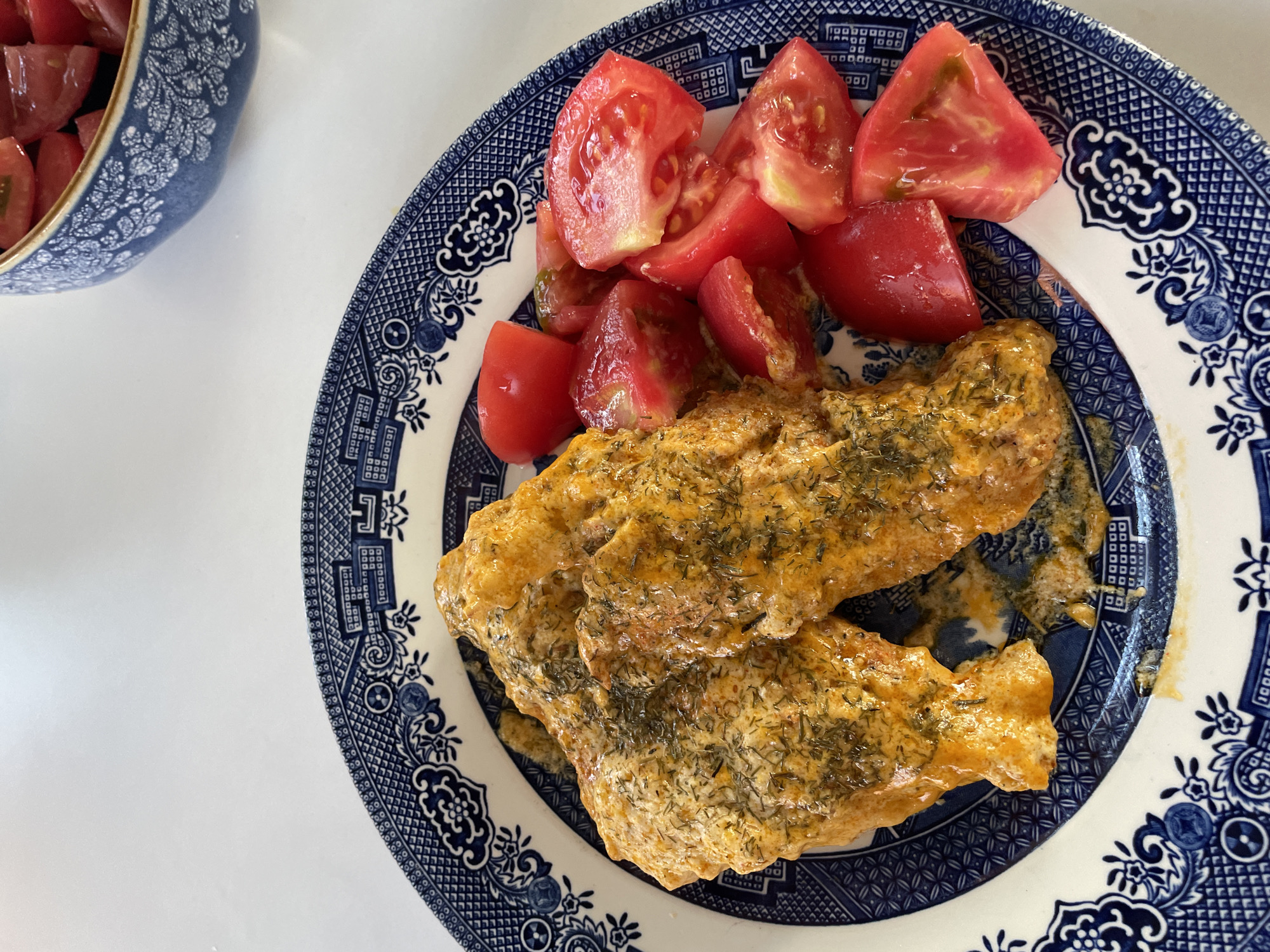 The image shows a close-up, overhead view of a blue and white patterned plate holding two pieces of food coated in a creamy, orange sauce and sprinkled with dill. To the right of the food are several chopped red tomatoes. In the background, a blue and white patterned bowl filled with more chopped tomatoes is visible. The setting appears to be a white table. The location is not specified as Skala Avdiron, Greece, but is a domestic indoor setting.