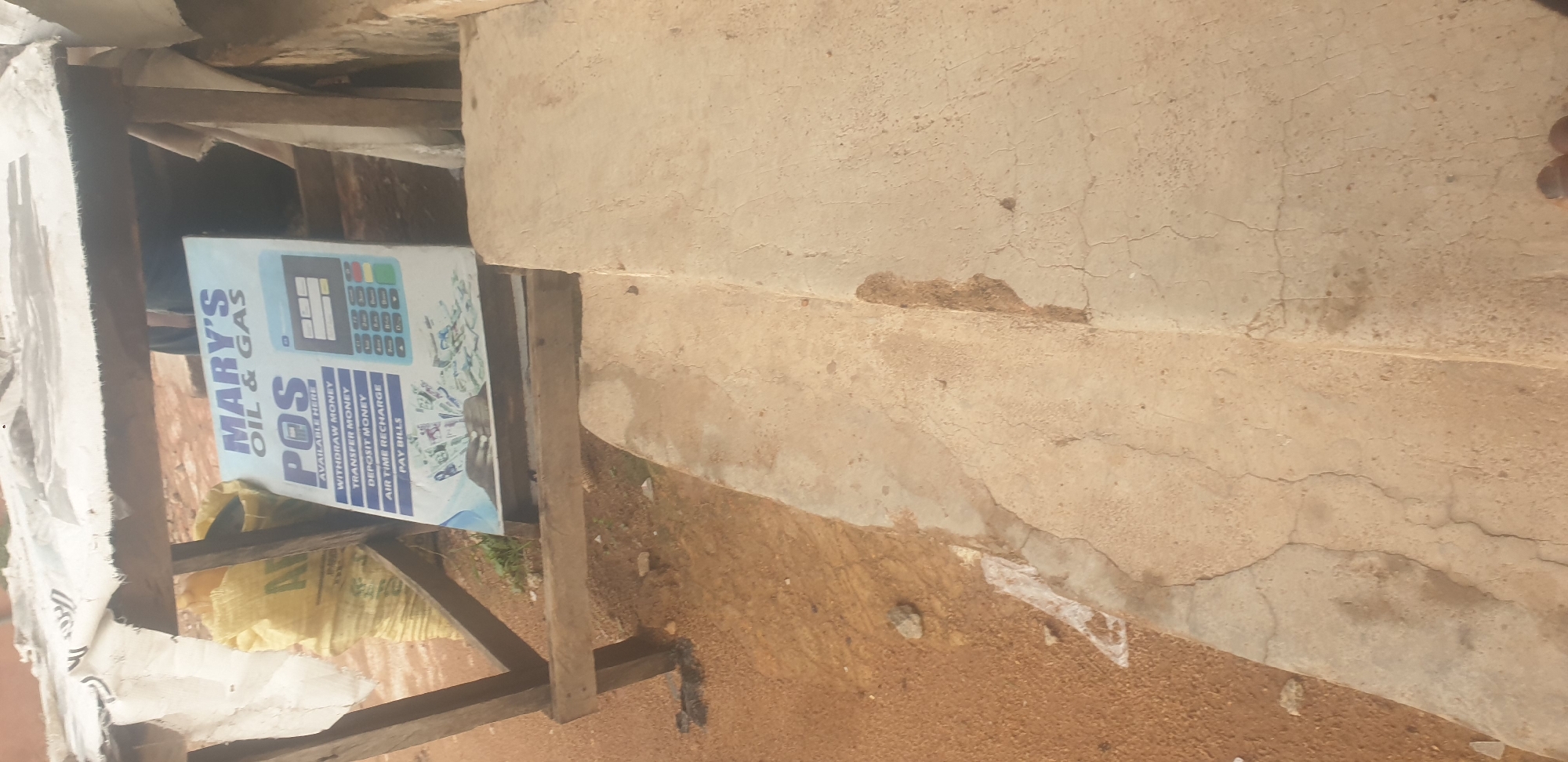 In the city of Jos, Nigeria, a POS (Point of Sale) advertising sign for "MARY'S OIL & GAS" is displayed at ground level, leaning against a wooden structure that appears to be a makeshift kiosk. The sign lists services offered: "WITHDRAW MONEY", "TRANSFER MONEY", "DEPOSIT MONEY", "AIR TIME RECHARGE", and "PAY BILLS". A graphic on the sign depicts hands holding various denominations of Nigerian Naira currency and a POS terminal. To the left of the sign, a large yellow sack is visible. The ground is covered in reddish-brown dirt, with scattered debris. A concrete step or platform is in the foreground, showing cracks and signs of wear. The overall scene suggests a roadside business operating in a semi-urban or rural setting.