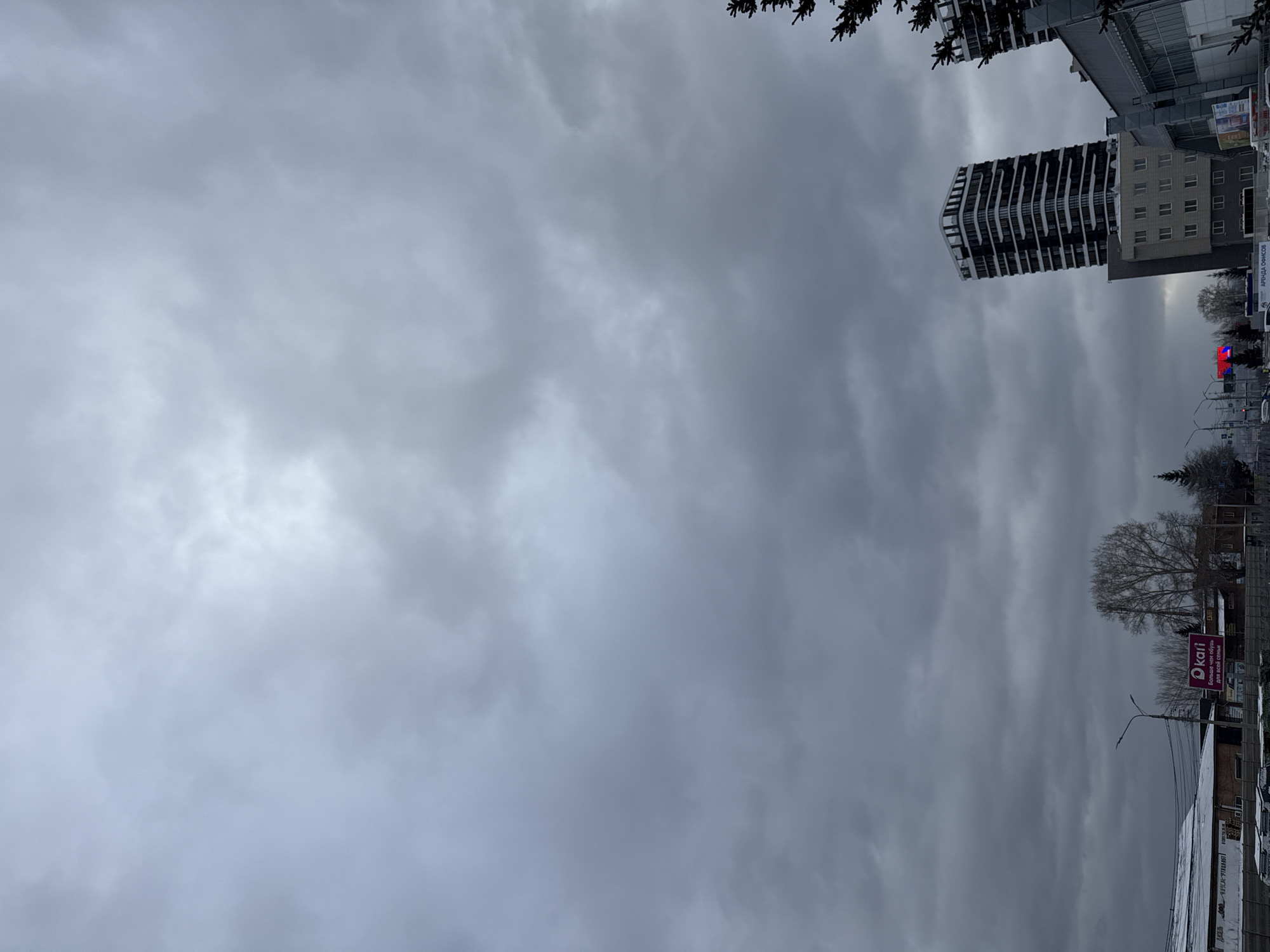 The image shows an overcast sky over what appears to be a street in Krasnoyarsk, Russia. A tall, modern building with a black and white facade is visible on the right side of the frame, suggesting an urban environment. A street-side sign is partially visible. Other buildings are present along the street, as well as trees. Cars are parked along the side of the street, and there is a streetlamp visible. The overall tone is somber and gray due to the heavy cloud cover.