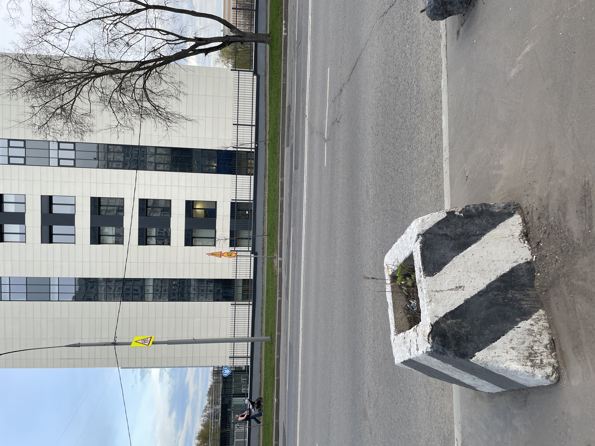 The image depicts a street scene in Khimki, Russia. A multi-story building with a white tiled facade and dark-framed windows stands behind a black metal fence. Bare tree branches are visible against the building. A road runs parallel to the building with a green median strip between the building and the road.
On the right side of the road, a concrete block painted with black and white diagonal stripes sits on the edge of the sidewalk. The block has a hollow top, containing some small plants and debris.
In the lower part of the image, a person is visible riding a bicycle on the sidewalk adjacent to the road. A yellow pedestrian crossing sign is mounted on a pole. Another street sign with numbers is also visible near the building. The sky is overcast with some patches of blue.