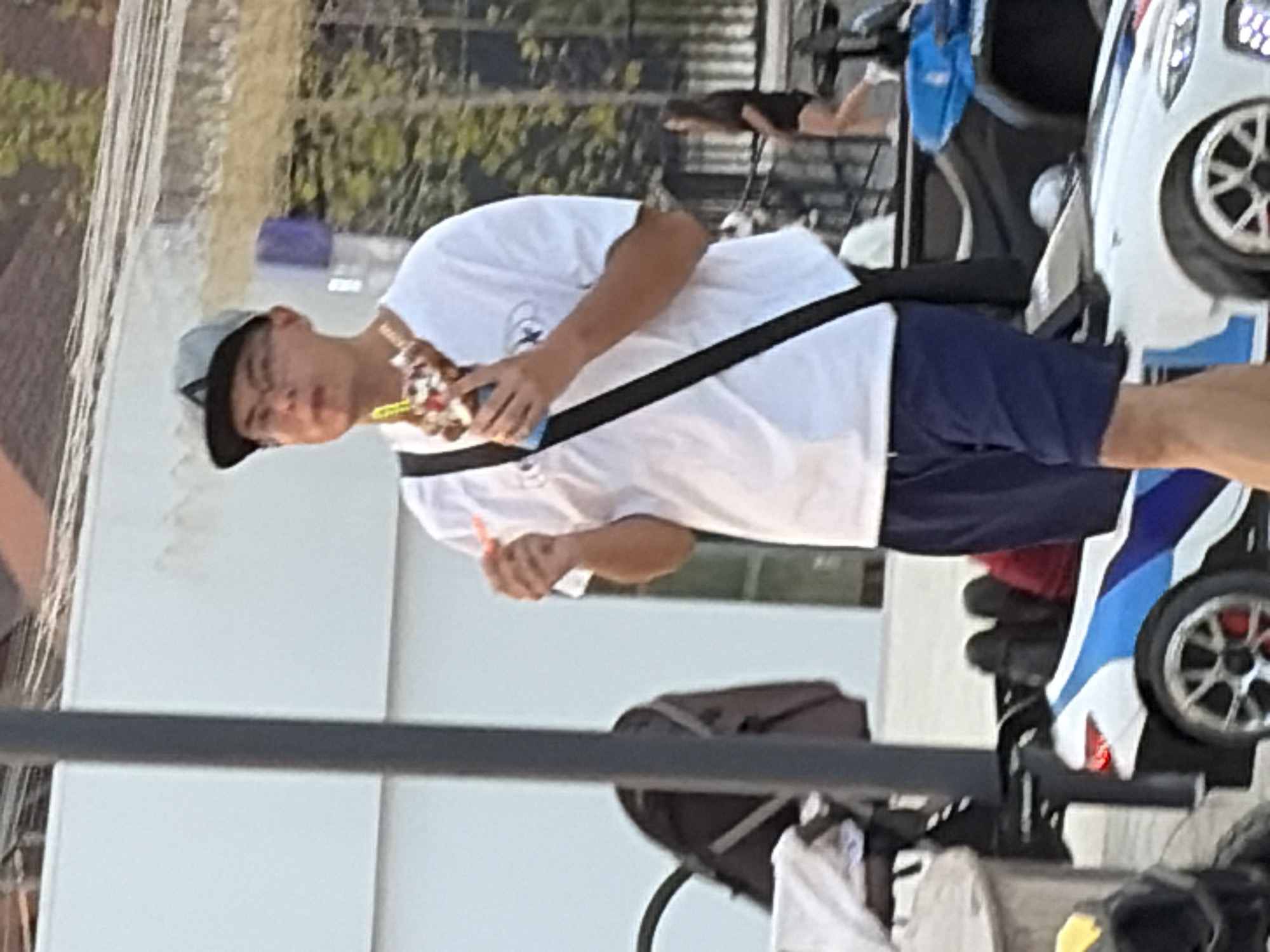 A young man wearing a white t-shirt, blue shorts, a grey baseball cap, and glasses is standing and eating a dessert that resembles a sundae. He is holding a spoon in his right hand, and the dessert is in his left hand. A black strap goes diagonally across his chest. In the foreground, to the left of the man, is a dark grey baby stroller with a white cloth draped over it. To the right of the stroller and in front of the man, is a white electric toy car with blue and black racing stripes and silver wheels. The car has red brake calipers. Behind the man, to the right, a dark-colored ATV is parked. Further in the background, a woman in a black shirt and shorts is walking, and trees and buildings are visible. The scene appears to be outdoors on a paved surface.