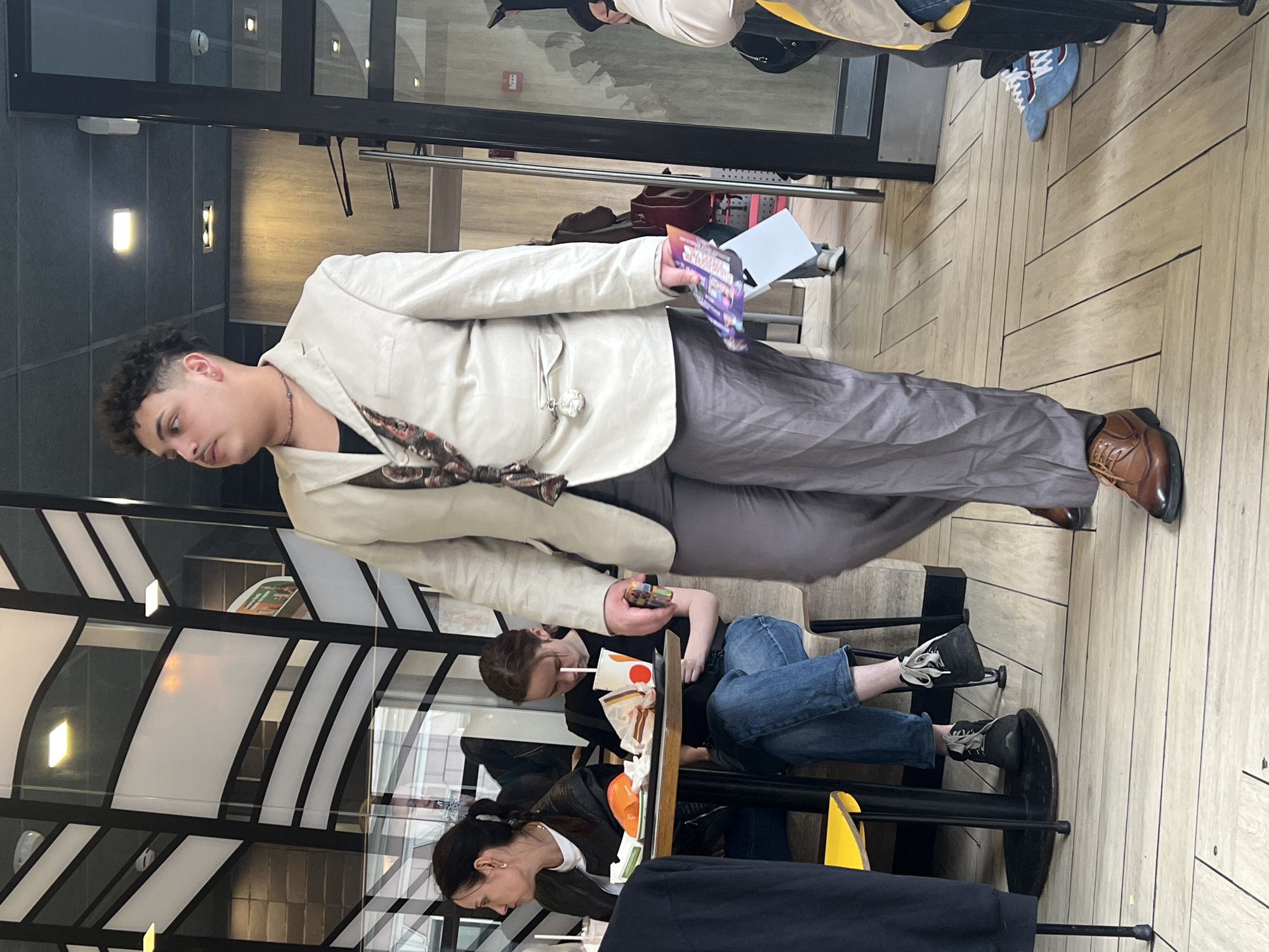 Inside a McDonald's restaurant, possibly in Saint Petersburg, Russia, due to the restaurant design which is similar to McDonald's locations there.
A male in an off-white blazer, gray pants, and brown dress shoes stands in the foreground. He is holding a leaflet in his left hand and a cell phone in his right. He is looking slightly to his left. He has curly dark hair.
Behind him, two women are seated at a table. One woman is wearing jeans, a black shirt, and black shoes, and is using her cell phone. The other woman is partially visible, wearing a black leather jacket and a white shirt.
Other patrons are visible at adjacent tables. The floor has a light wood-grain pattern. The McDonald's restaurant has black and white architectural details.