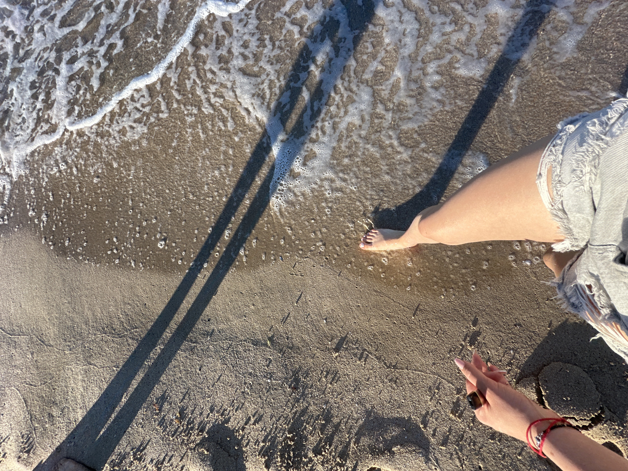 An overhead shot of a person's legs and feet on a sandy beach at the edge of the sea. The person is wearing ripped denim shorts. The right foot is bare and partially submerged in the foamy sea water. The left hand, adorned with a ring and a red bracelet, is resting on the sand. The sun casts a long shadow across the sand from an unseen object. The scene is set in Primorsko, Bulgaria.