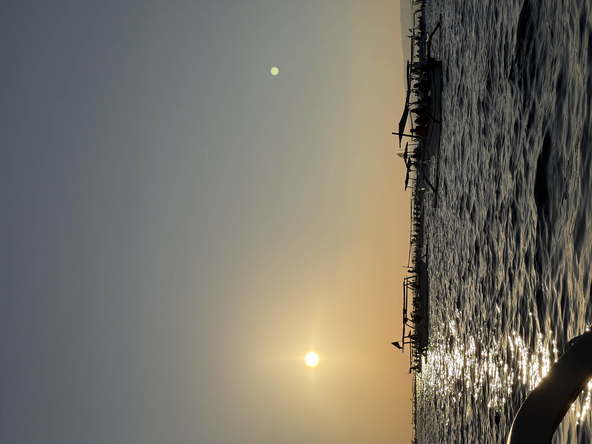 A photograph captures a sunset over the ocean in Buleleng, Indonesia. Several silhouetted traditional Indonesian outrigger boats are visible on the dark water, some carrying passengers. The sun, a bright orb, is low in the sky and casts a glittering reflection on the choppy waves. The sky transitions from a pale yellow near the horizon to a dusky blue at the top of the frame. The overall mood is serene and peaceful.