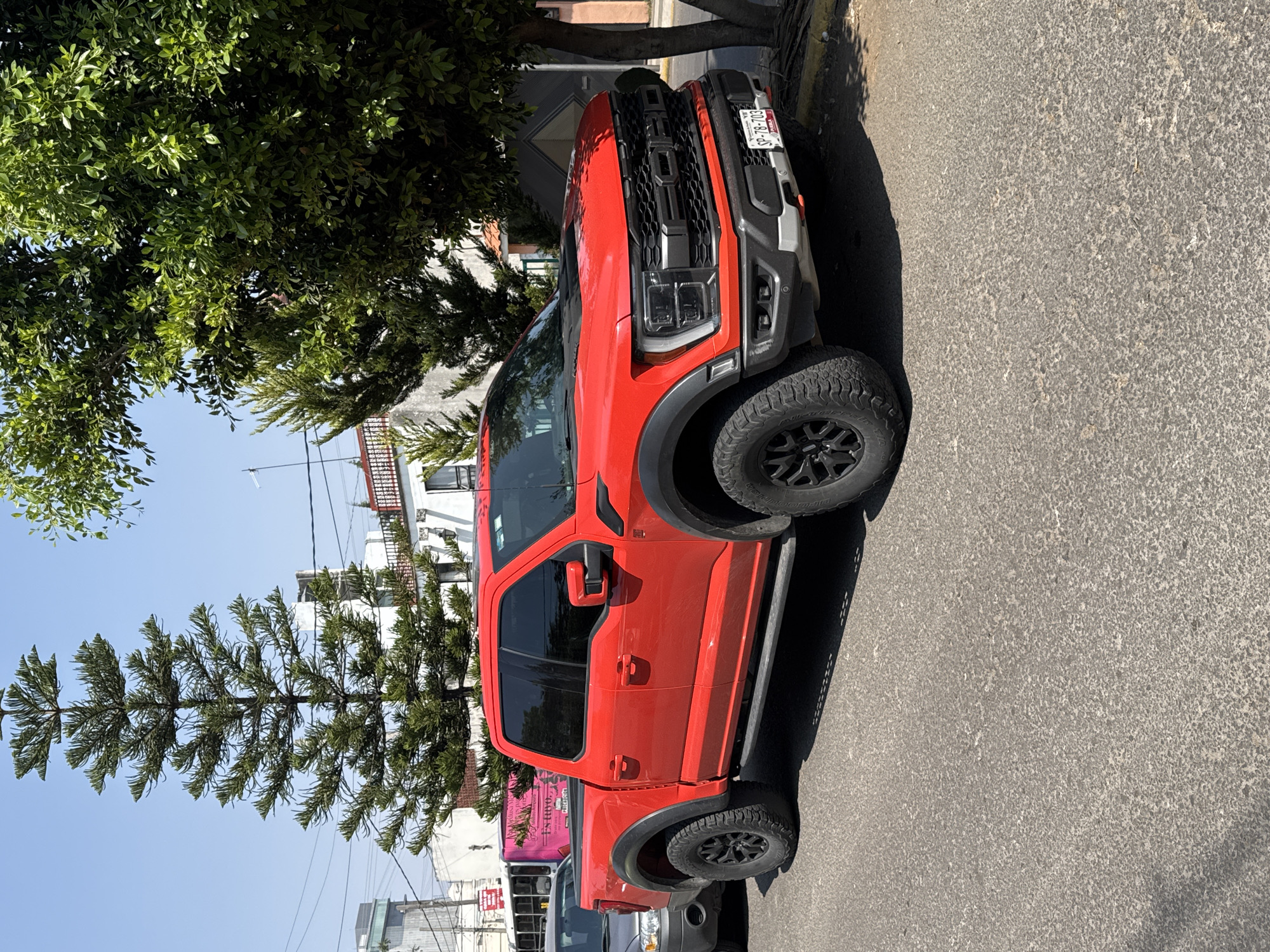 An eye-level outdoor shot shows a red Ford Raptor pickup truck parked on the side of a street in the city of Puebla, Mexico. The truck is parked parallel to the curb on an asphalt road. The front passenger side of the truck faces the camera. The front license plate reads "SP-78-703." The wheels have black rims. A large tree hangs over the front of the truck. In the background are buildings, some white with multi-story architecture. A second, gray truck can be seen behind the red one, along with a pink bus. The sky is blue.