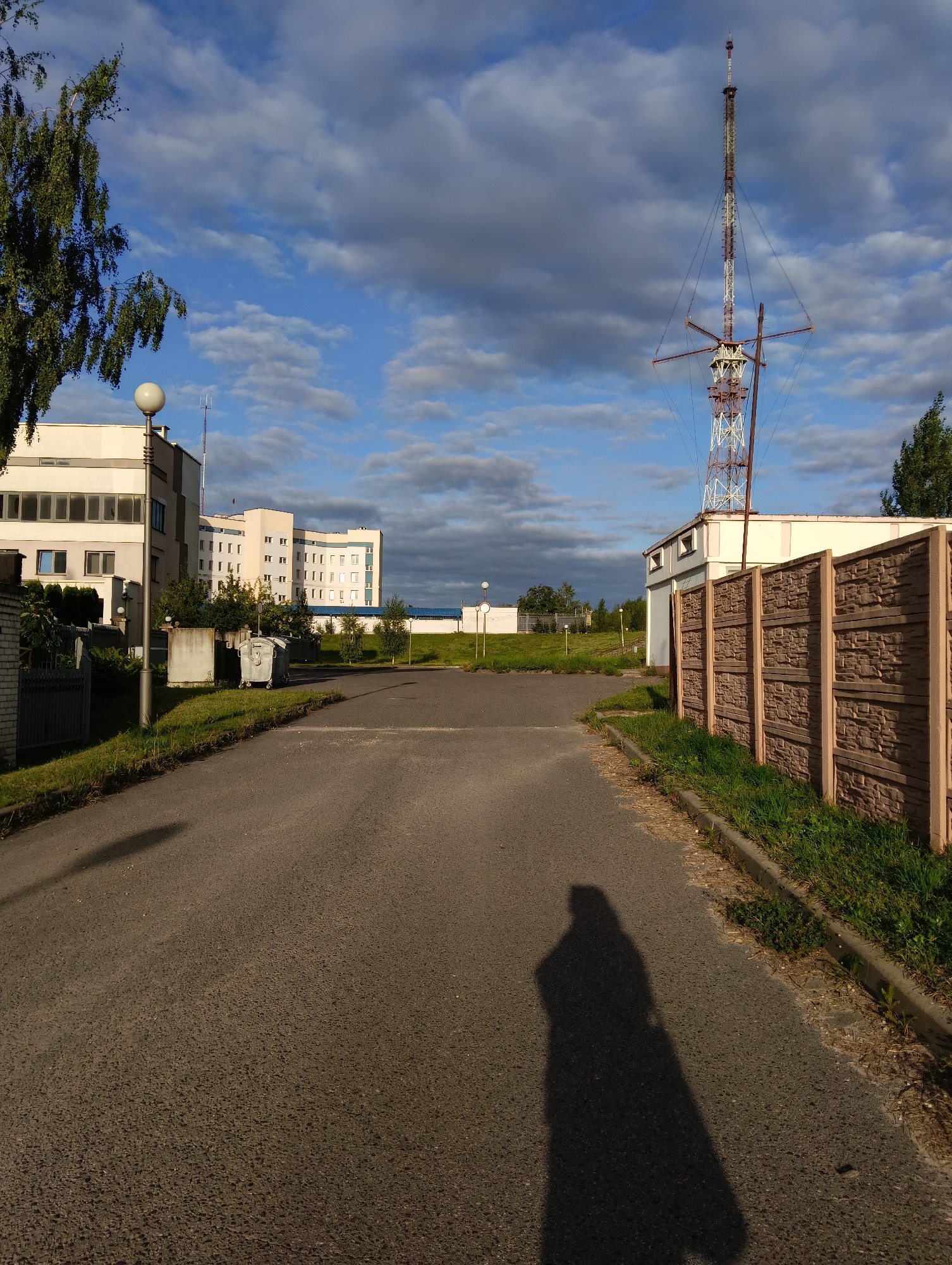 The image depicts an asphalt road leading towards several buildings and a tall radio tower. On the left side of the road, there is a white building with multiple windows, a lamppost, and a fence. In the background, a larger, multi-story white building with many windows is visible. On the right side of the road, a decorative concrete fence separates the road from a grassy area. Behind the fence, a building with a light-colored facade and a row of windows can be seen. The prominent radio tower, with its lattice structure and antenna at the top, stands against a sky filled with dramatic clouds. The shadow of a person, suggesting the photographer, is cast on the road in the foreground. The scene appears to be in an industrial or administrative area, likely on the outskirts of the city.