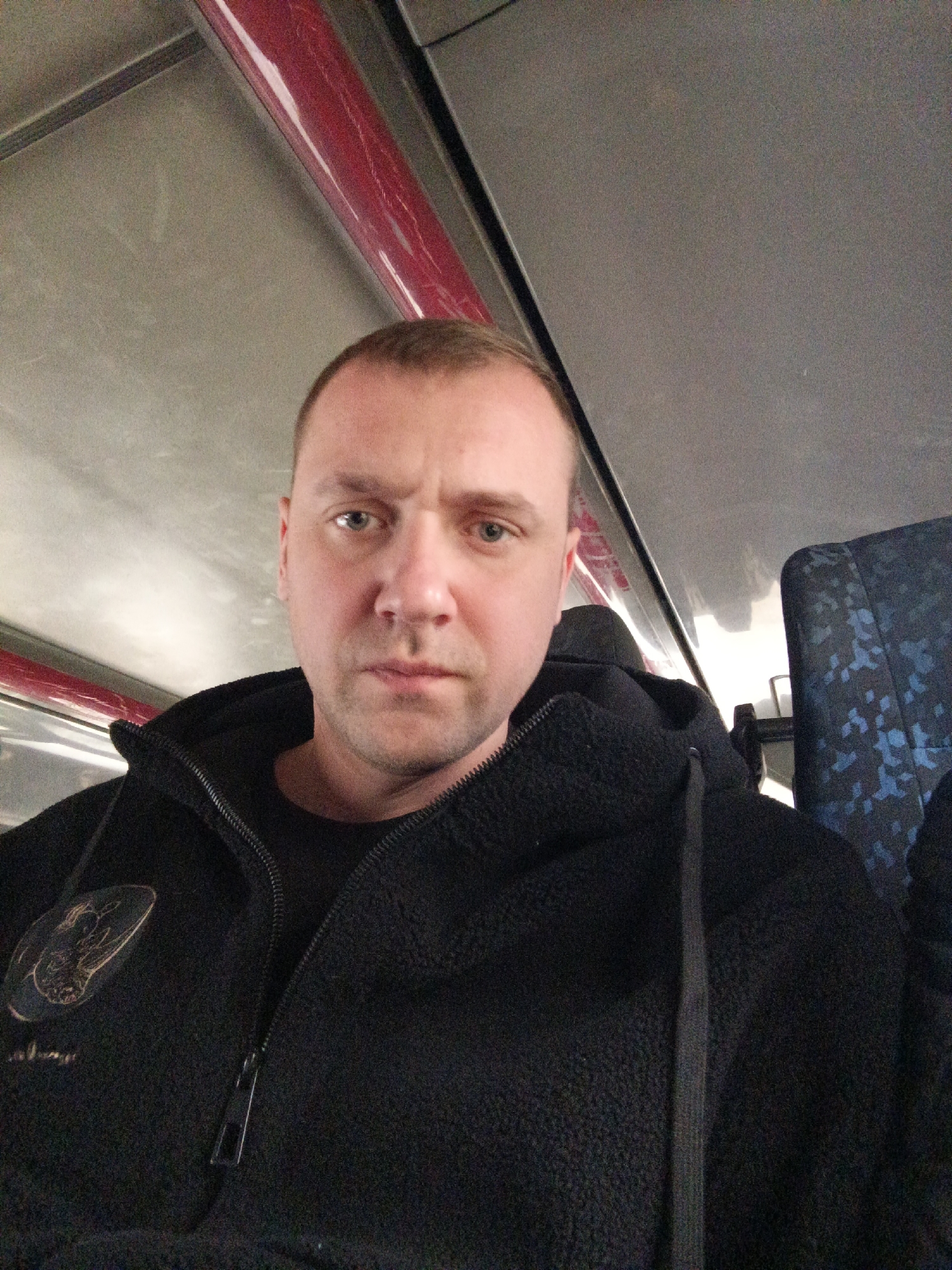 A man, appearing to be in his 30s or 40s, is captured in a head-and-shoulders selfie shot, looking directly at the camera with a neutral to slightly serious expression. He has short, light-colored hair and light-colored eyes.
He is wearing a black, textured fleece hoodie or jacket, partially zipped up over a dark t-shirt. On the left side of his chest, there is a light-colored emblem or patch, featuring an animal design (possibly a lion) within an oval, with unreadable text beneath it. The drawstring of his hoodie is visible on the right side of his chest.
The setting appears to be the interior of a public transport vehicle, likely a train or bus, consistent with travel in Klin, Russia. The ceiling is light-colored, possibly grey or white, and a prominent red bar or pipe runs along the upper left portion of the interior. Reflections of light sources are visible on the ceiling. To the man's right, a passenger seat is visible, upholstered in dark fabric with a repeating light blue or white pattern. The lighting suggests it is daytime.