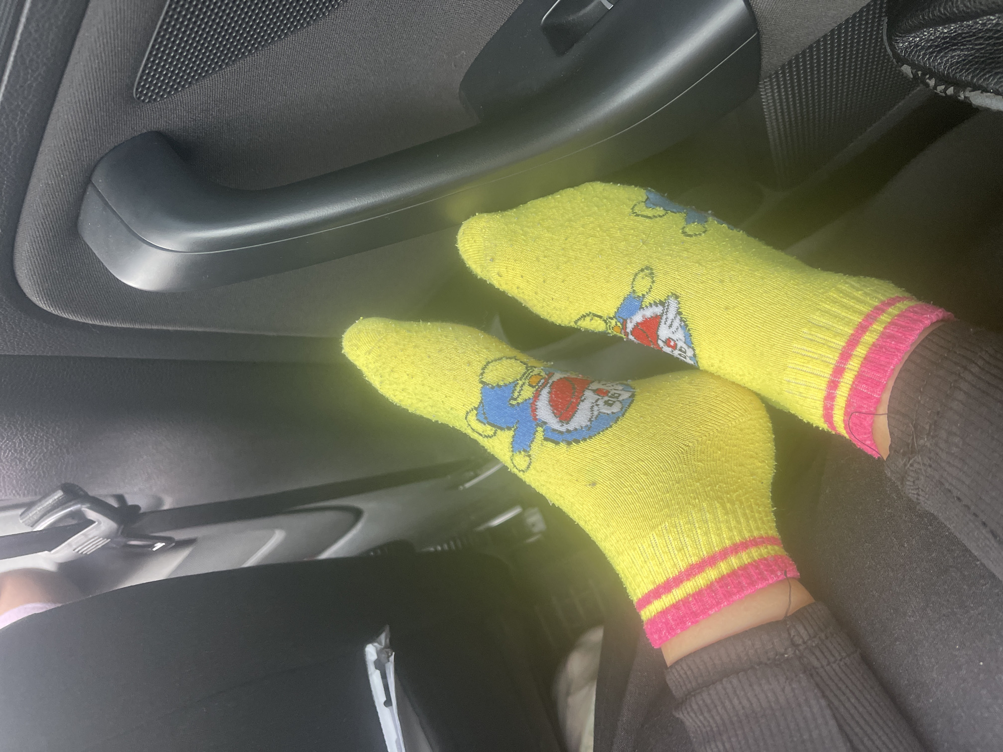 The image shows a close-up view of two feet wearing bright yellow socks with pink and yellow stripes around the cuffs. Both socks feature a cartoon character, appearing to be Doraemon, an anthropomorphic robotic cat. The socks are positioned on the floor of a vehicle, with the dark interior of the car, including a door handle and part of a seatbelt, visible in the background. The setting is likely a car parked in or near the village of Horozhuli (Гожули), Ukraine, as indicated by the provided location.