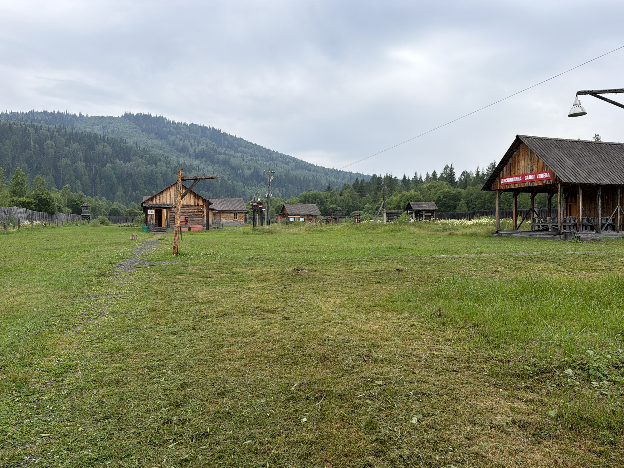 The image depicts an outdoor scene in a rural setting, likely a village or an agricultural compound, under an overcast sky. In the foreground, a large expanse of mown grass covers the ground, with patches of longer grass and some scattered wildflowers on the right. A narrow gravel path winds through the grass towards a cluster of wooden buildings.
To the left, a rustic wooden fence encloses an area, and a small wooden guard tower or observation post stands within it. Several log cabins and smaller wooden structures are visible, appearing to be part of a settlement. One of these buildings has a sign with Russian text in red.
In the midground, more wooden structures are scattered across the grassy field. A prominent wooden building with a large open-fronted section, possibly a covered market or workshop, is situated on the right side of the image. A large, cone-shaped lamp is suspended from a metal pole above this structure.
The background is dominated by a dense, green, tree-covered mountain range, indicative of a forested, mountainous region. The overall impression is of a remote, possibly historical or traditional settlement, nestled within a natural landscape. The location is identified as Ust'-Kabyrza, Russia.