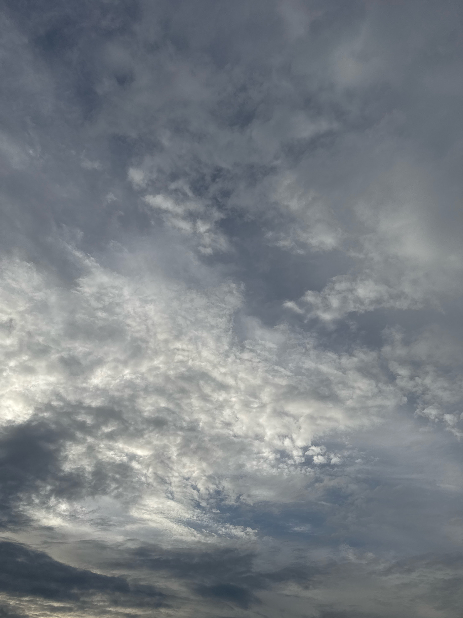 The image depicts a cloudy sky over what appears to be a rural or suburban area, possibly the Бараўлянскі сельскі Савет in Belarus. The clouds are varied, with some appearing dense and dark, suggesting an overcast or approaching storm, while others are lighter and more scattered, allowing glimpses of sunlight to filter through. The overall color palette is dominated by grays and whites, with hints of blue sky visible in some areas. There are no people, structures, or distinct objects visible in the image, focusing solely on the atmospheric conditions. The lighting is diffused, creating a somewhat dramatic and moody ambiance.