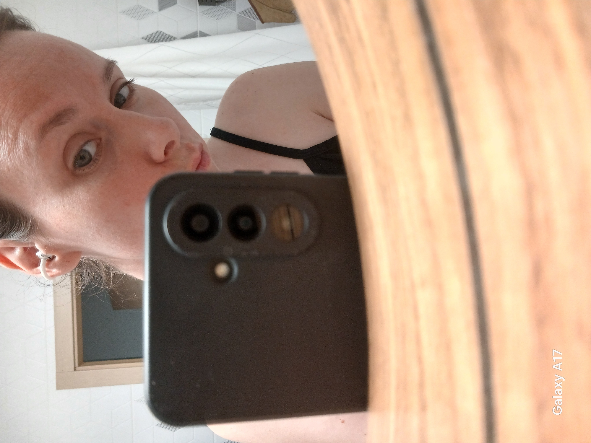 A woman is taking a selfie in a bathroom mirror. The phone is blocking a large portion of the view. The background shows a tiled wall and a window frame.