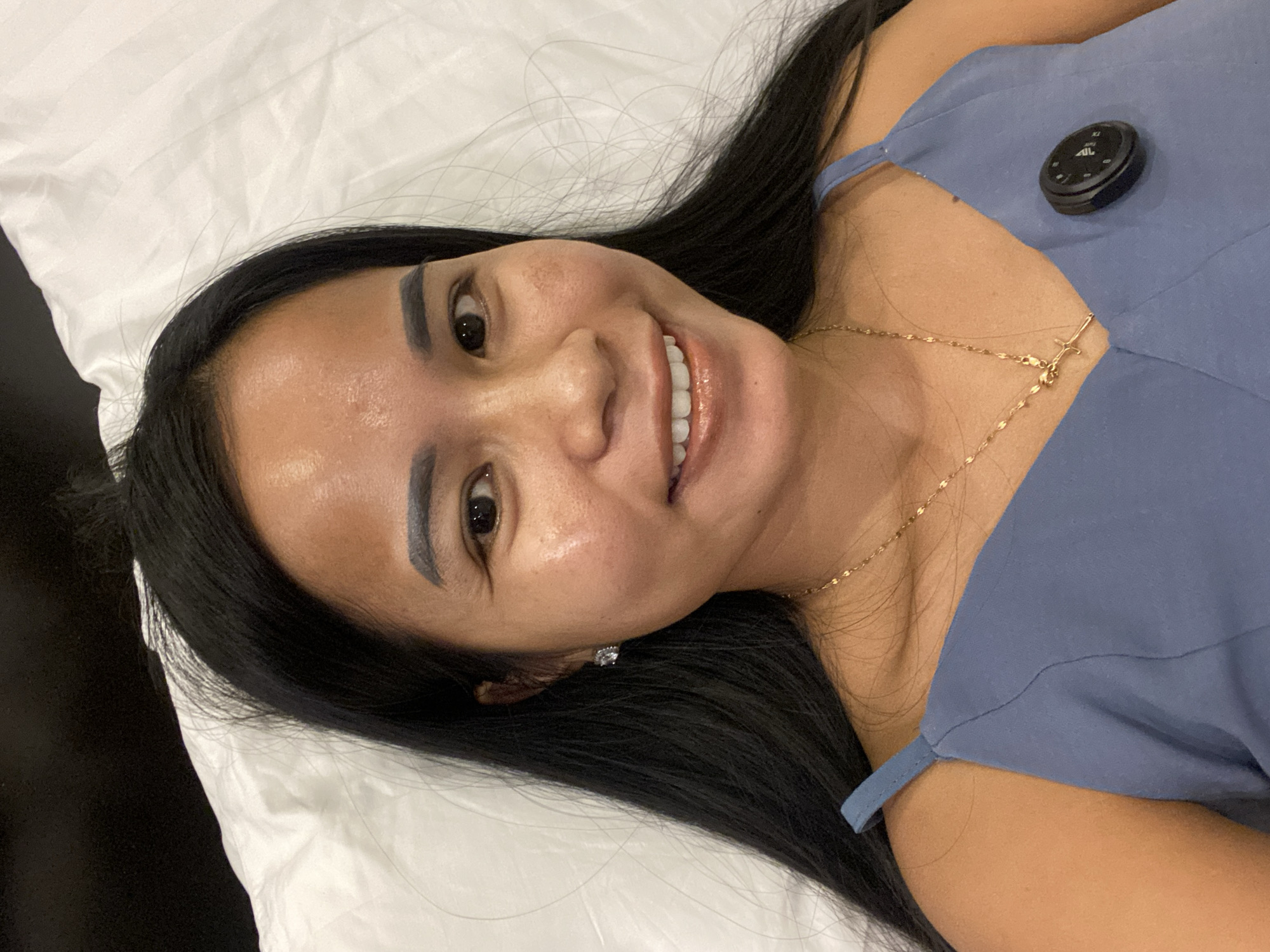 A woman with long black hair is lying on a white pillow. She is smiling and wearing a light blue top and a gold necklace with a cross pendant. A small black device is clipped to her top. She appears to be in an indoor setting. The location is North Kuta, Indonesia.