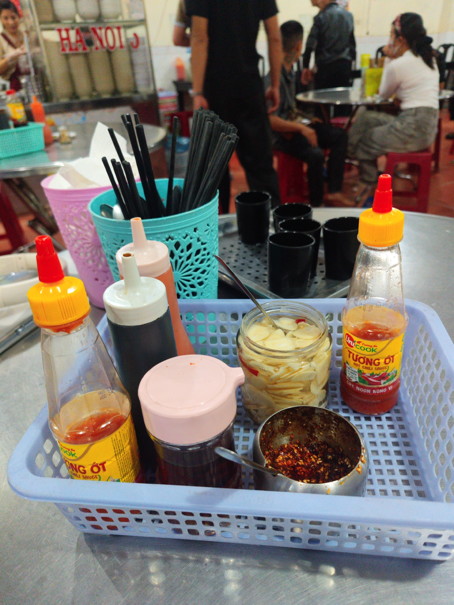 The image shows a table setting in a Vietnamese restaurant, likely in Hanoi, Vietnam, with condiments and dining utensils. The table is a metal surface, possibly stainless steel.
A light blue plastic basket holds several condiments. There are two bottles of "Tuong Ot" hot chili sauce with yellow and red caps. There are also two other bottles with pink and white caps, presumed to contain other sauces. A jar of sliced pickled garlic is visible, along with a small metal container holding chili flakes in oil. A spoon is present in the jar of garlic and the container of chili flakes.
Behind the basket are containers holding utensils. A pink container and a turquoise container hold chopsticks, while a metal tray contains small, black cups. The chopsticks are black.
In the background, there are several other tables with patrons. A sign with the word "HANOI" is visible, and several people are walking around. Some are sitting and eating. The general ambiance suggests a bustling, casual dining environment.