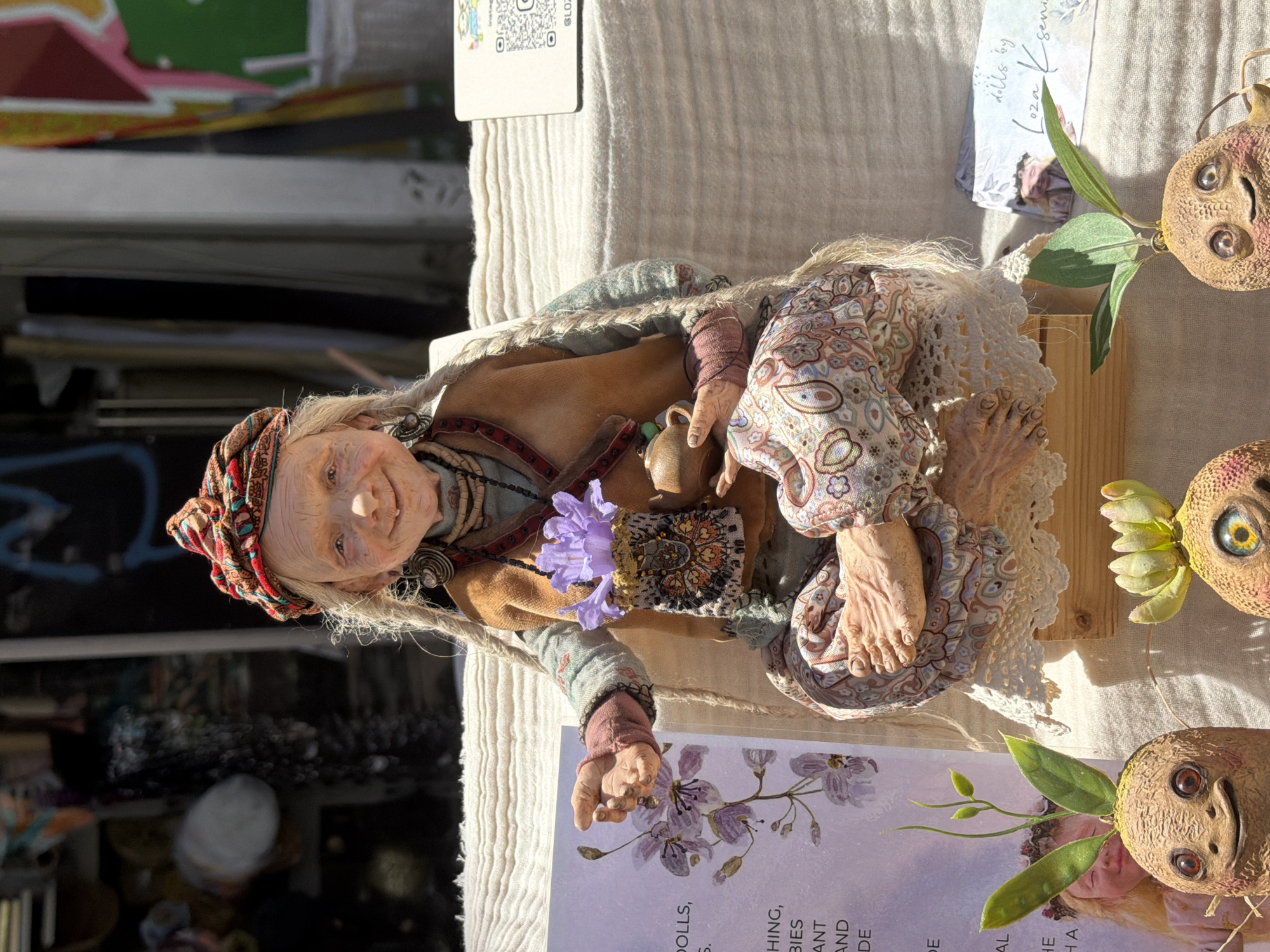 The image captures a handmade doll, depicting a wrinkled woman with fair hair under a red and orange patterned headscarf. She wears a tan outfit with a decorated vest, and patterned pants, and holds a small brown jug. The doll sits on a wooden platform covered with a white crocheted fabric. To the right are two ceramic mandrake-like objects with leaves. The doll and objects are placed on a table with a white linen tablecloth, with promotional cards visible. The text on one of the cards reads "DOLLS...DOLLS BY Loza Ksenia." The overall setting suggests an artisan market or craft fair. Tel-Aviv, Israel can't be conclusively confirmed in this image.