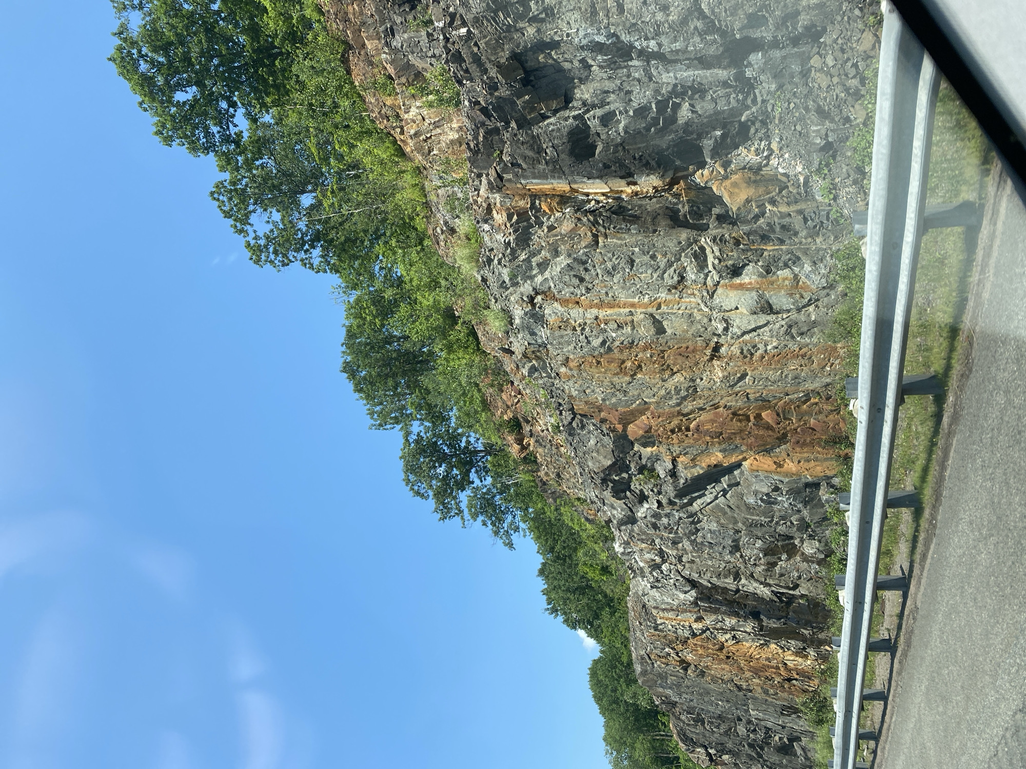 The image shows a roadside rock face in Pointe-au-Chêne, Canada. The rock face is steep and jagged, with streaks of orange and brown iron staining. Trees and vegetation grow along the top and sides of the rock face. A metal guardrail separates the road from the rock face. The road appears to be paved and is visible in the foreground. The sky is blue with a few wispy clouds. The overall impression is of a natural landscape that has been modified for road construction.