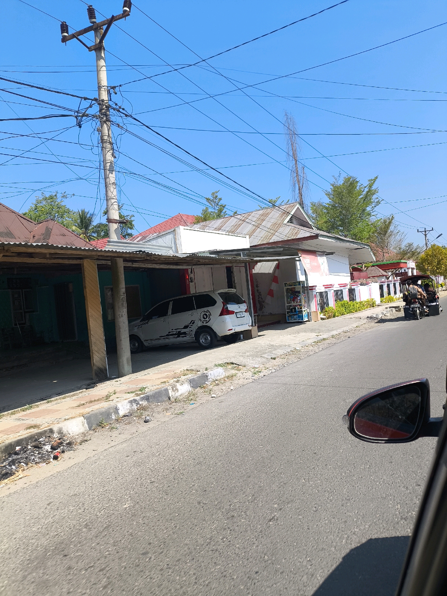 This is a photograph taken from a vehicle, looking forward and slightly to the right. The scene is an urban or semi-urban environment in Tilamuta, Indonesia, under a clear blue sky. The road surface is asphalt, with a concrete curb and a drainage ditch on the right side. The ditch contains some scattered debris and a red bottle.
On the left side of the road, a row of low-rise buildings is visible. The first building has a teal-colored facade and a covered porch supported by wooden beams. A white multi-purpose vehicle (MPV) with black and grey graphics is parked under this porch. Further down the road, there is a longer building with a white and red facade, featuring a canopy extending over a sidewalk. This building appears to be a small shop or convenience store, with shelves visible through a window displaying various products. Green foliage lines the front of this building.
Further down the street, a motorized tricycle, commonly known as a "bajaj" or "becak motor," is traveling away from the camera. It carries three passengers. Beyond this, another building with a red roof is visible, and a utility pole with a complex network of electrical wires stands prominently in the mid-ground. Trees with green leaves are scattered throughout the scene, some taller and more mature, others smaller. The overall impression is of a sunny day in a tropical or subtropical region. The camera captures a reflection of what appears to be another vehicle in the side-view mirror on the far right of the frame.