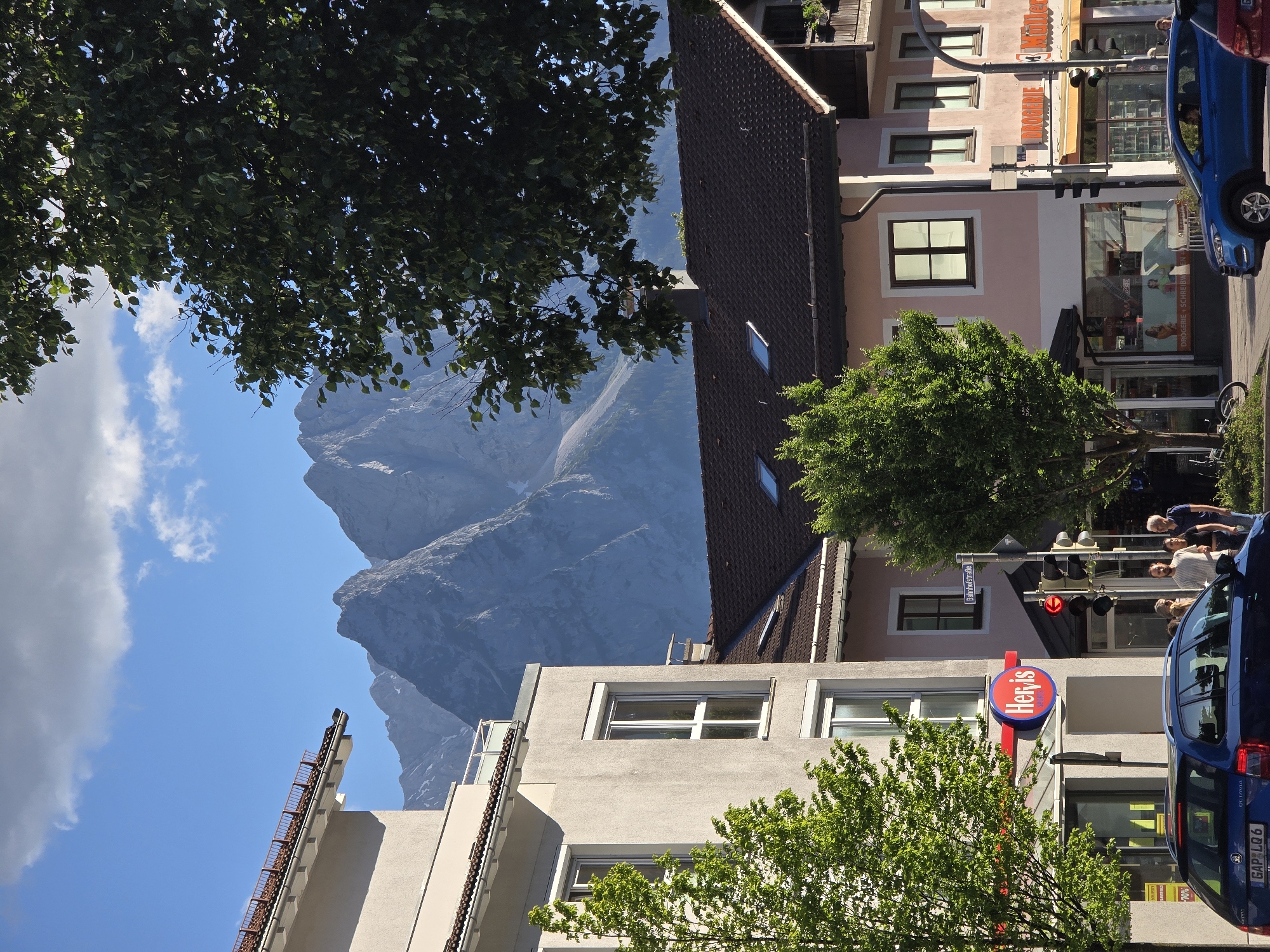 The image depicts a street scene in Garmisch-Partenkirchen, Germany, with a backdrop of majestic mountains. In the foreground, a blue car is parked on the street, with its license plate visible. To the left of the car, a building with a red and white sign that reads "Her is SPORTS" is situated. Several trees with lush green foliage are scattered throughout the scene, some in pots and others planted directly in the ground. In the middle ground, more buildings line the street, some with shops and others with residential apartments. A few people are visible walking on the sidewalk, engaged in conversation or looking at shop displays. The buildings feature a mix of architectural styles, with some having red tiled roofs and others flat roofs. The sky is a clear blue, with a few fluffy white clouds scattered across it. The mountains in the background are rugged and snow-capped, adding a dramatic touch to the otherwise ordinary street scene. The overall impression is one of a charming European town on a bright, sunny day.
