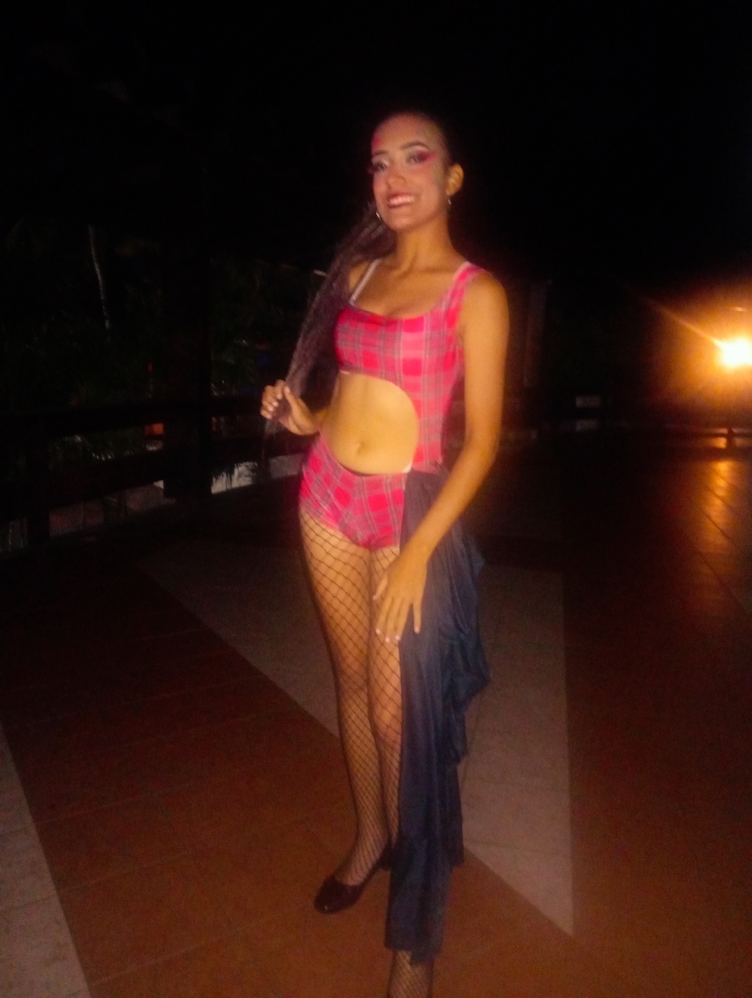 A woman stands on a tiled terrace in Playa El Agua, Venezuela. She wears a pink plaid swimsuit with a cut-out at the abdomen, fishnet stockings, and black shoes. She holds her hair in her left hand. A black cloth hangs from her right side. Behind her is a dark railing with vegetation and a lit area.