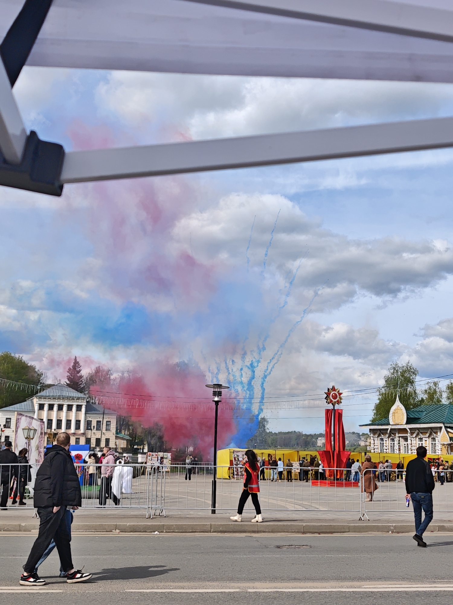 The image captures a scene in Uglich, Russia, featuring red and blue smoke in the background. In the foreground, there's a street with two individuals walking on the left and one on the right. A crowd of people is visible behind a metal fence in the central part of the image. Behind them is a red monument with a star on top, flanked by buildings with traditional Russian architecture. The sky is partly cloudy with blue patches, and a portion of a tent structure is visible in the top-left corner of the frame.