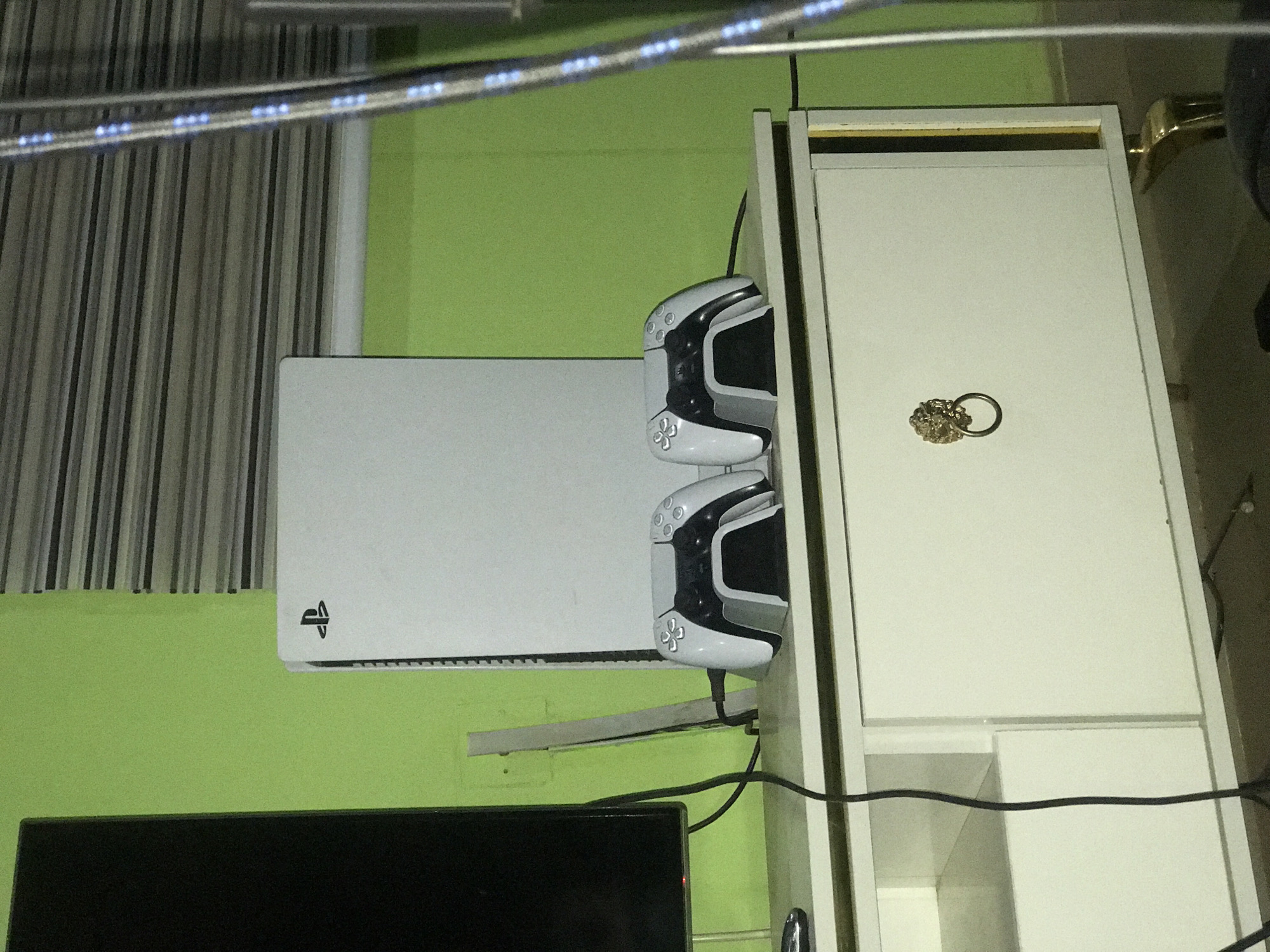 The image displays an indoor gaming setup, prominently featuring a white PlayStation 5 (PS5) console mounted horizontally on a light green wall. The console's distinctive 'PS' logo is visible on its left side. Adjacent to the PS5, two white and black PlayStation DualSense controllers are neatly stacked in what appears to be a charging or storage dock, also affixed to the wall or an adjacent structure.
To the right of the gaming console, a white cabinet or shelving unit is present. On its top surface, a metallic key ring adorned with an ornate, golden-colored embellishment, possibly a stylized animal head or intricate design, rests. In the bottom left corner of the frame, a section of a black television screen is visible. Above the console, a window with vertical or venetian blinds is partially seen. A string of small blue LED lights traverses the upper part of the image, suggesting decorative lighting. Various black cables are interconnected with the gaming equipment.
The scene depicts a contemporary home entertainment arrangement, likely within a personal room in Abuja, Nigeria, indicating a space dedicated to gaming. The time of day is not explicitly clear, but the presence of LED lights might suggest evening hours or an aesthetic choice.