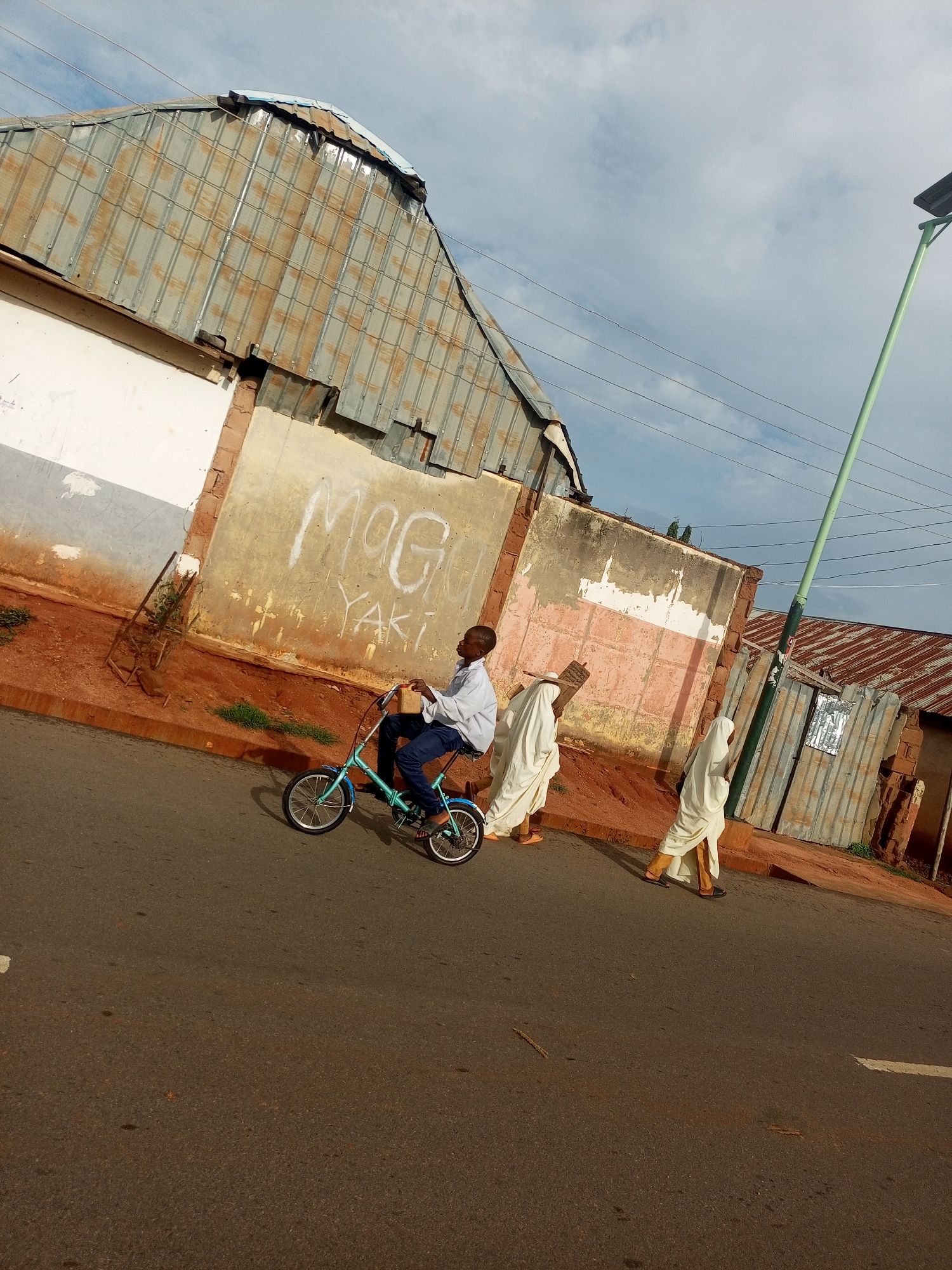 The image captures a street scene in Tempure, Nigeria, under a cloudy sky. In the foreground, a young boy rides a small blue bicycle on an asphalt road. He is wearing a white long-sleeved shirt and blue trousers. To his right, two individuals walk away from the camera, both draped in flowing white garments. The building in the background features a corrugated metal roof and a light-colored wall with graffiti. A tall, green lamppost stands on the right side of the frame. The environment appears somewhat weathered, with visible signs of wear on the buildings and a reddish-brown dirt area bordering the road.